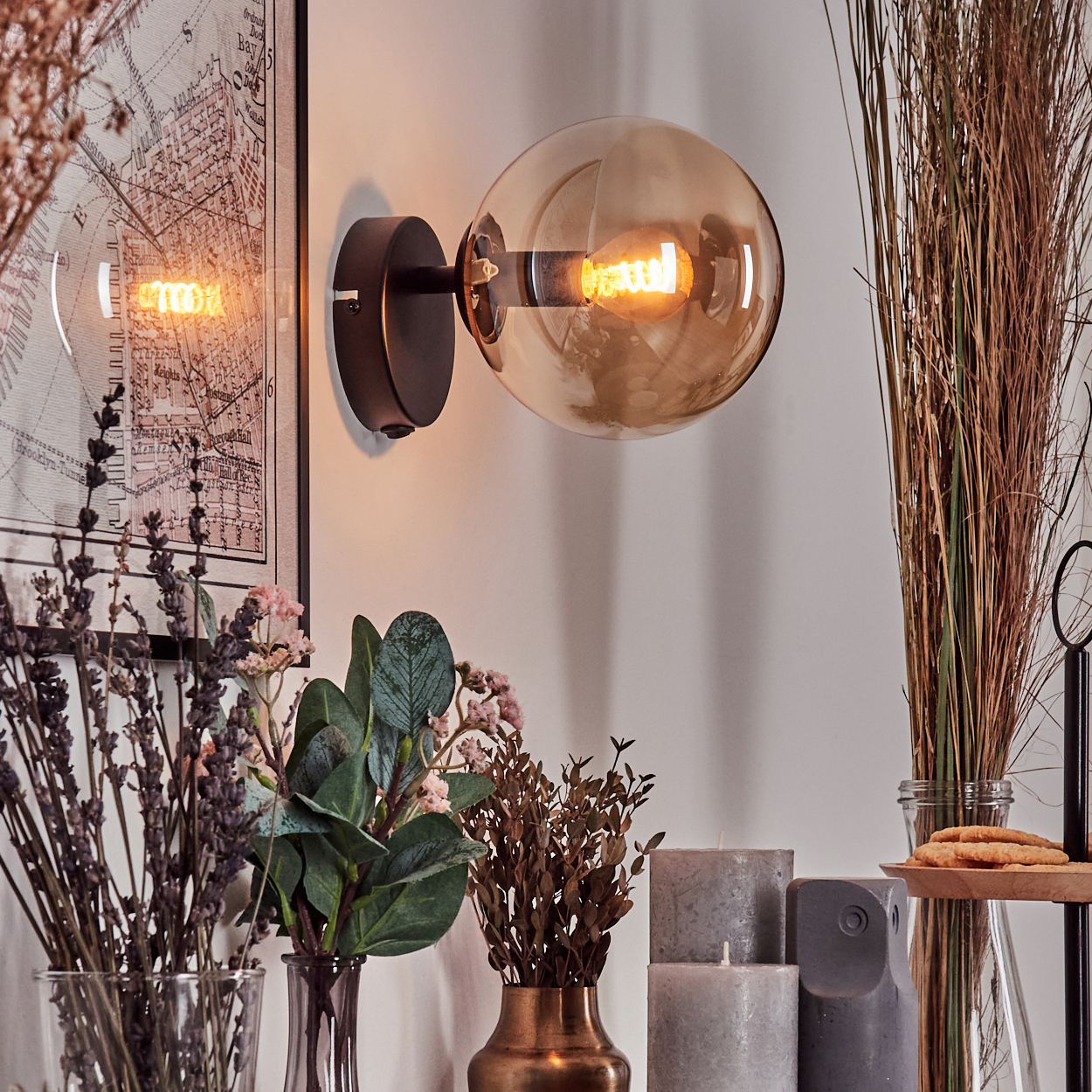 Eine Wandlampe mit Glasschirm leuchtet, über Vasen mit Blumen und Kerzen. Eine Karte hängt an der Wand.