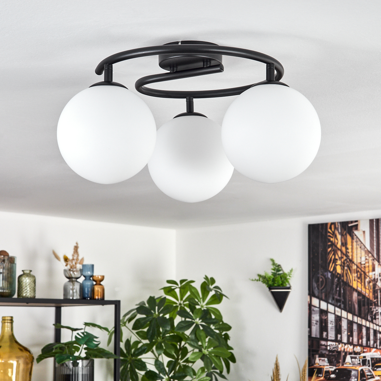 Moderne Deckenlampe mit drei weißen Kugelleuchten. Die Lampe ist schwarz, mit einem dekorativen Wirbel. Pflanzen und Kunstwerke sind unten sichtbar.