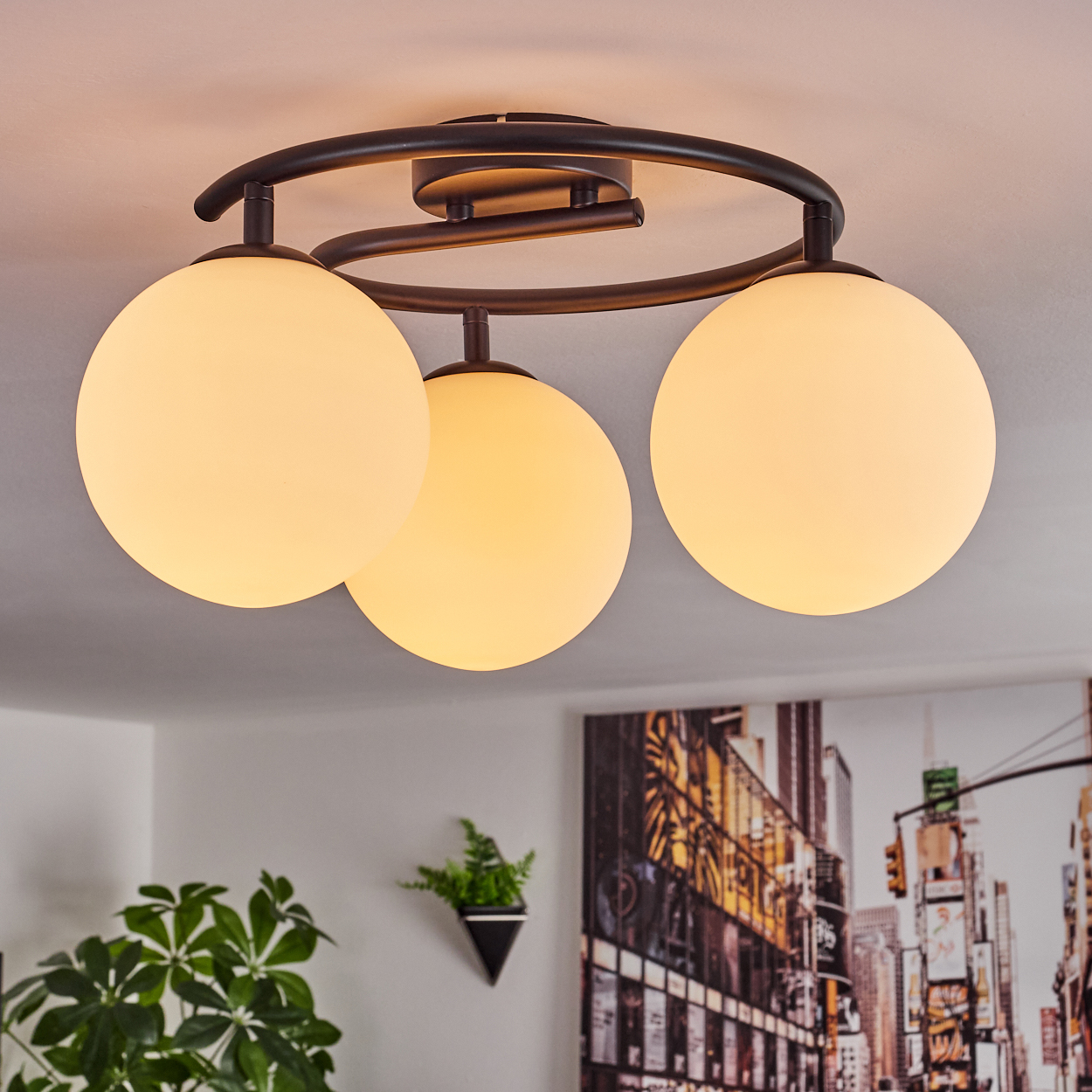 Deckenleuchte mit drei kugelförmigen Lampen. Die Leuchte ist schwarz und die Lampen sind weiß. Im Hintergrund ist ein Stadtbildgemälde zu sehen.