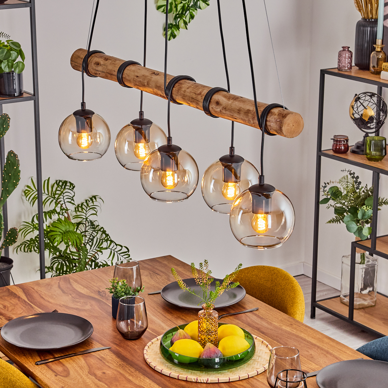 Esszimmertisch mit Holztisch, Hängelampen, Tellern, Gläsern und einer Obstplatte. Im Hintergrund befinden sich Pflanzen und Regale.
