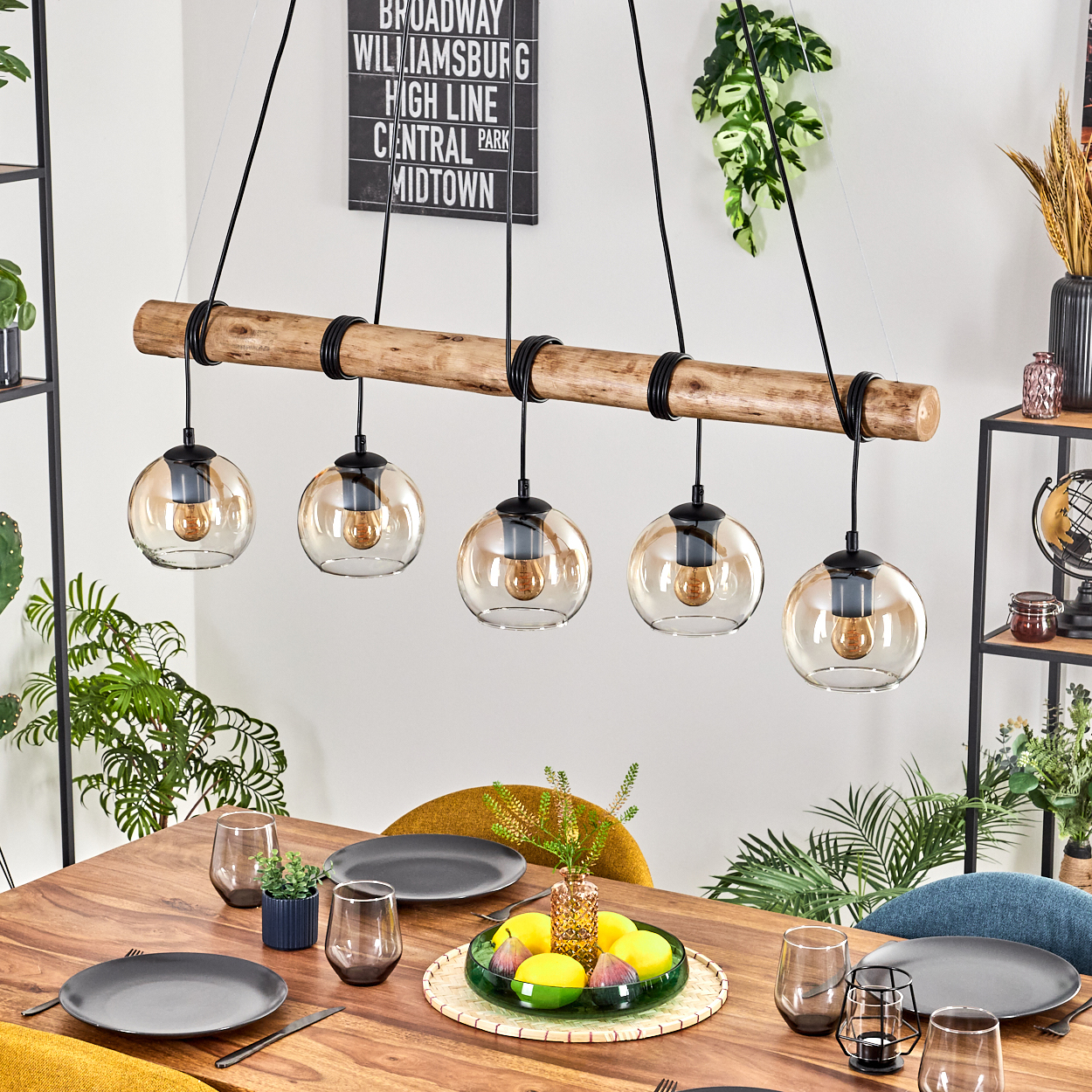 Ein Esstisch mit Tellern, Gläsern und einem Mittelstück aus Früchten. Darüber hängt ein Holzbalken mit fünf Pendelleuchten.