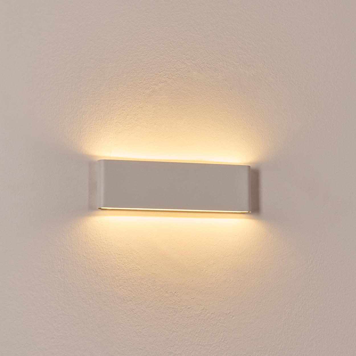 Eine rechteckige, graue Wandleuchte strahlt warmes Licht auf eine strukturierte weiße Wand. Das Licht breitet sich nach oben und unten aus.