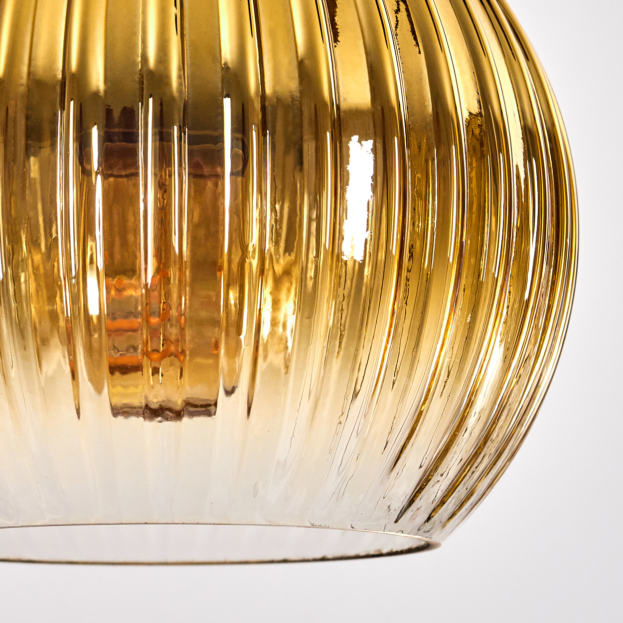 Nahaufnahme eines goldenen, gerippten Glaslampenschirms. Im Inneren ist eine Glühbirne zu sehen. Der Hintergrund ist weiß.