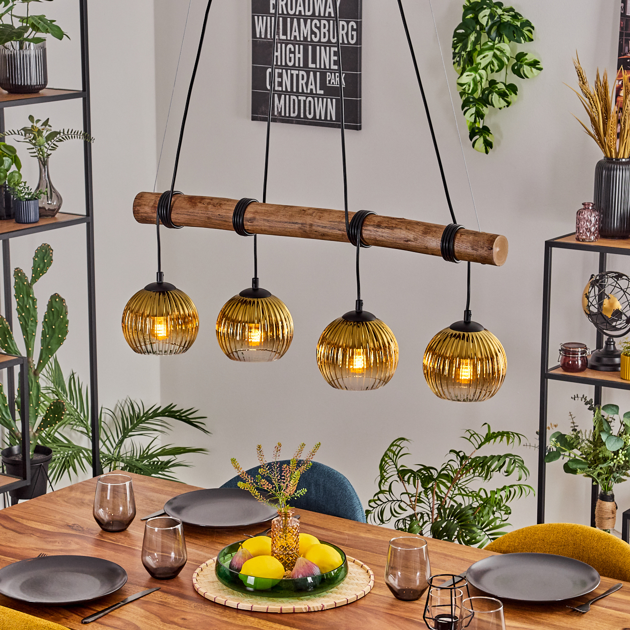 Esszimmer mit Holztisch, goldenen Pendelleuchten und Pflanzen. Tisch gedeckt mit Tellern, Gläsern und einer Obstschale.