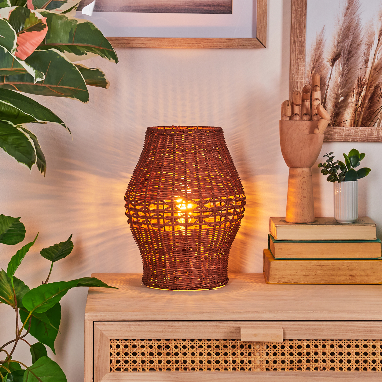 Eine beleuchtete, braune, gewebte Lampe steht auf einem Holzschrank. Pflanzen und gerahmte Kunstwerke sind im Hintergrund.