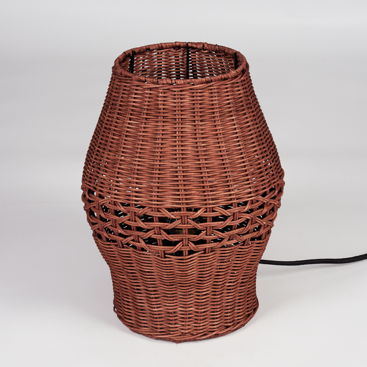 Eine braune Korblampe steht auf einer weißen Oberfläche. Die Lampe hat eine zylindrische Form mit einem Webmuster und ein schwarzes Kabel.