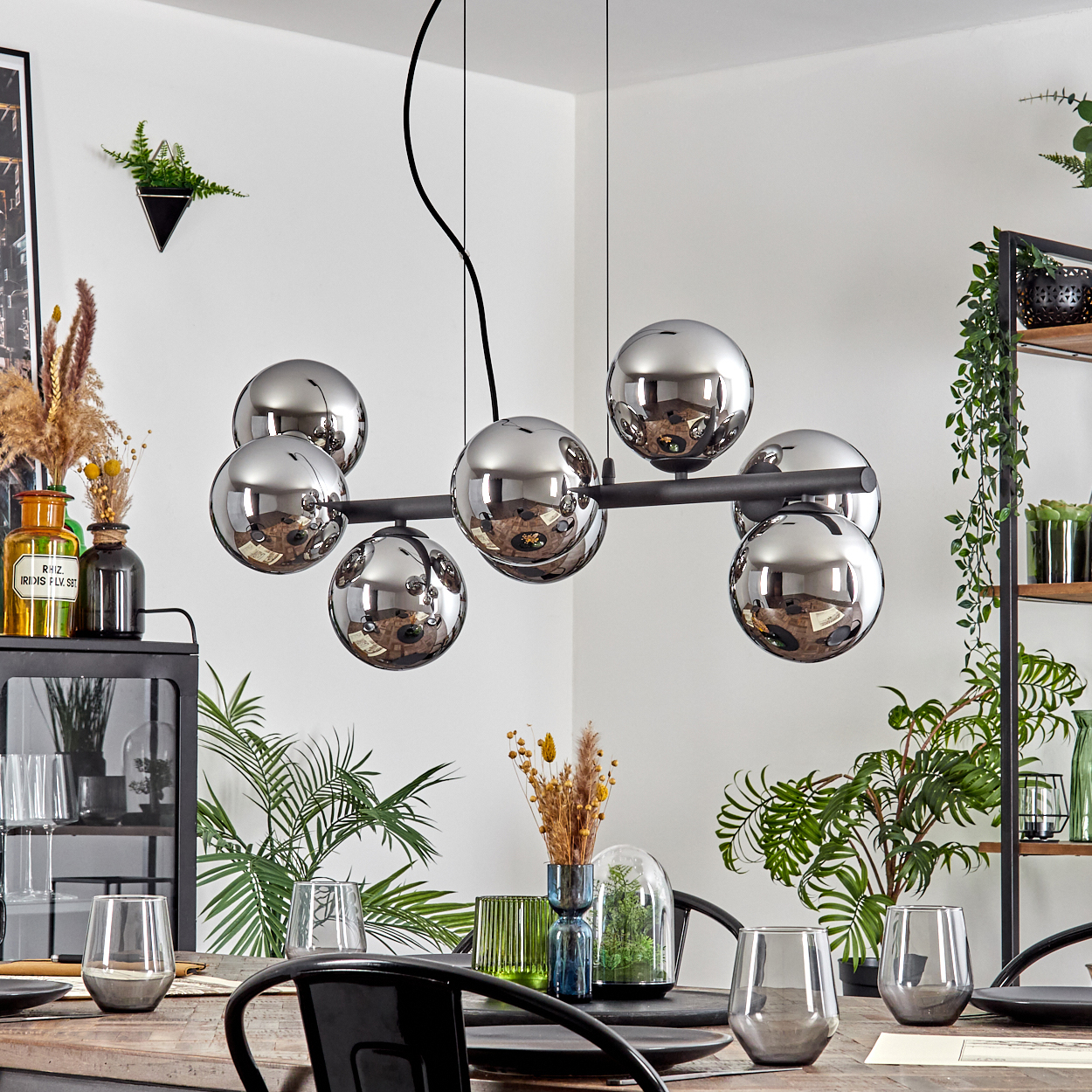 Ein Esszimmer mit moderner Leuchte mit Chromkugeln. Tisch mit Gläsern und Dekor, Pflanzen im Hintergrund gedeckt.