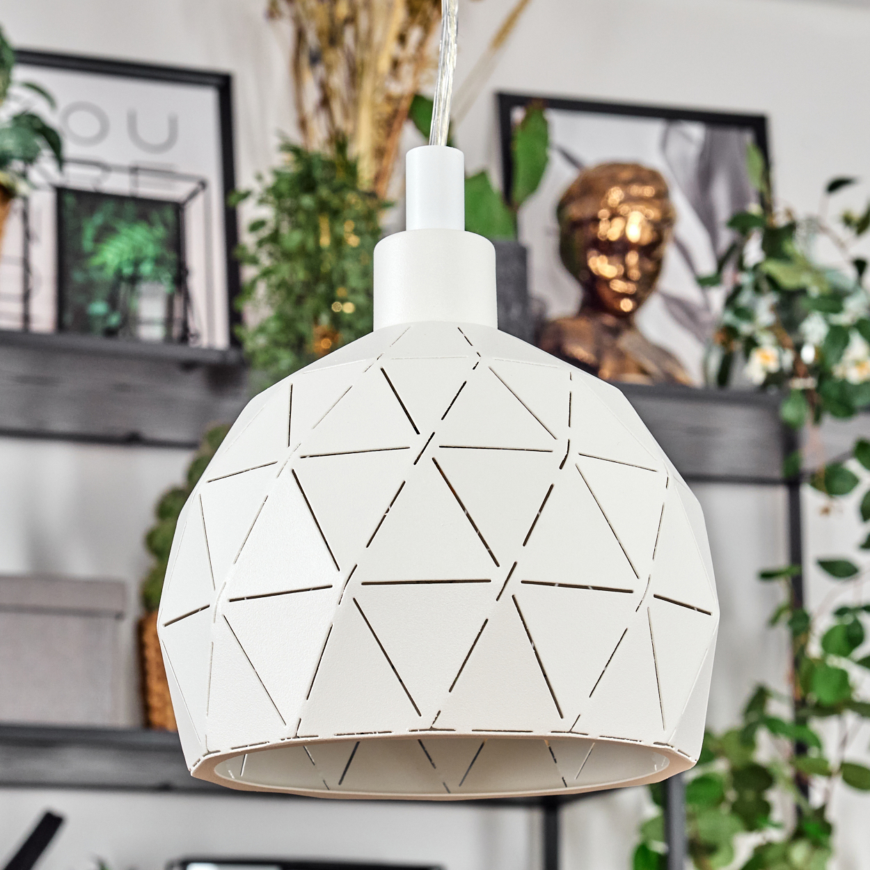 Eine weiße geometrische Pendelleuchte hängt vor einem Regal mit Pflanzen, Rahmen und einer Bronzebüste.
