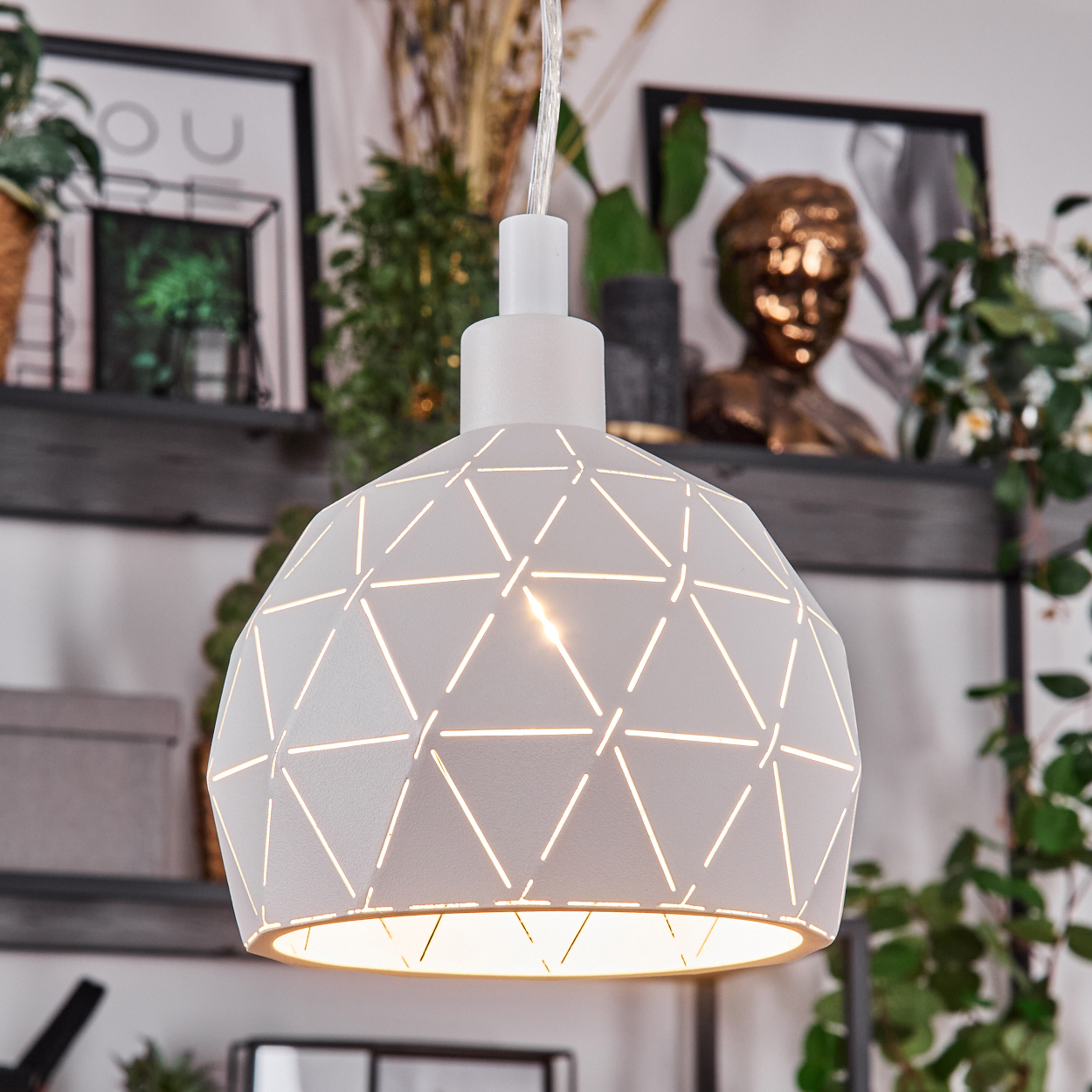 Eine weiße geometrische Pendelleuchte hängt und leuchtet. Regale halten Pflanzen und Kunst im unscharfen Hintergrund. Ein gemütliches, beleuchtetes Interieur.