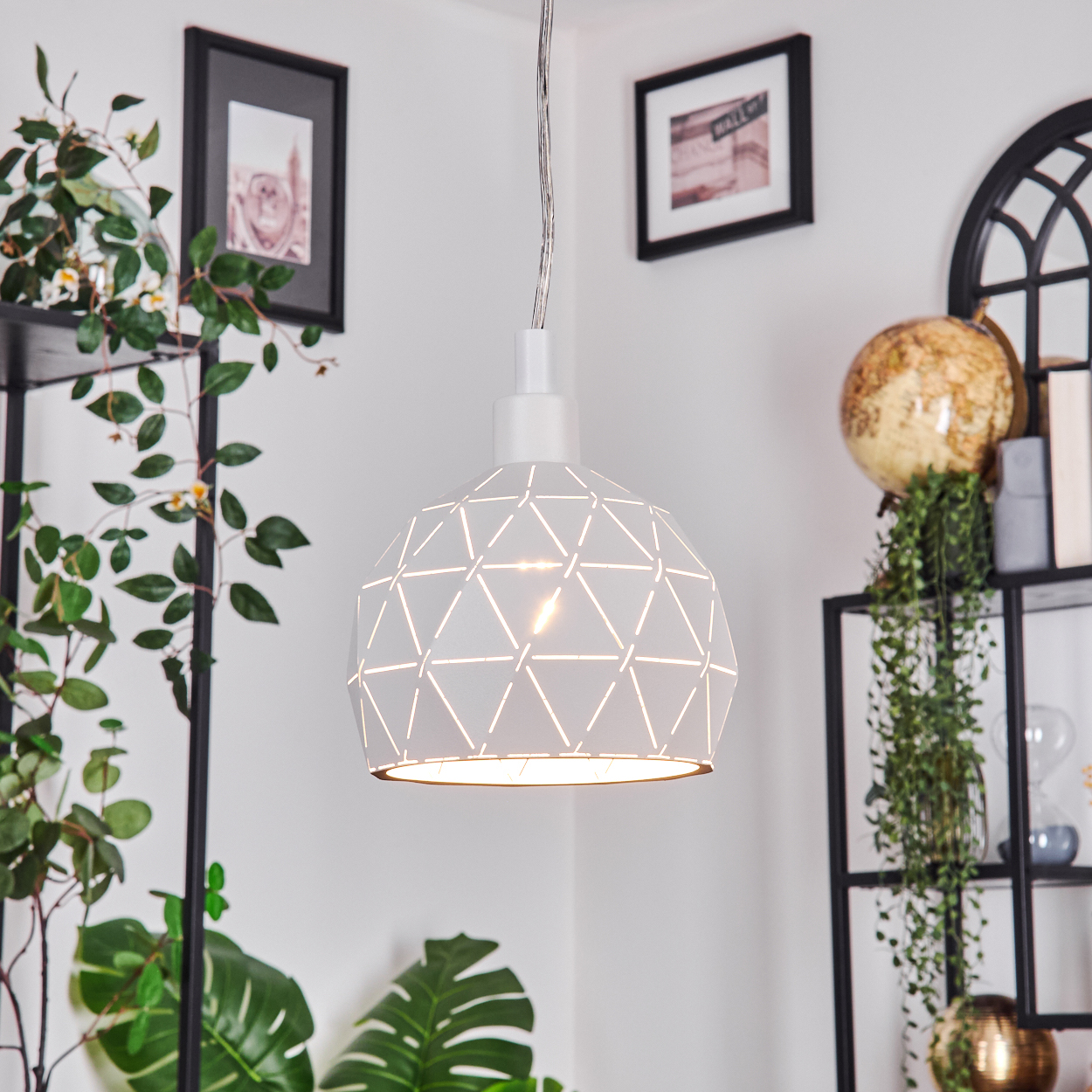 Eine weiße, geometrische Pendelleuchte hängt vor einer weißen Wand. Bilder, Pflanzen und ein Globus befinden sich auf den Regalen.