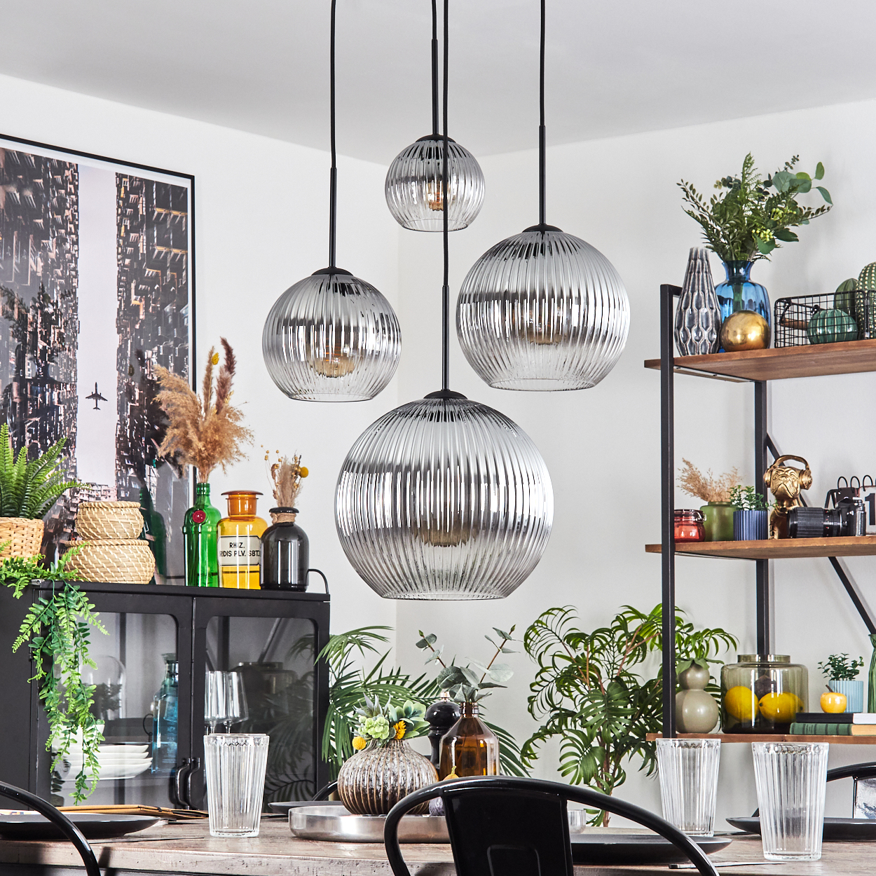 Esszimmer mit Hängelampen, Tisch, Pflanzen und Dekorationsartikeln. Silberne Lampenschirme, schwarze Akzente und ein Stadtbild.
