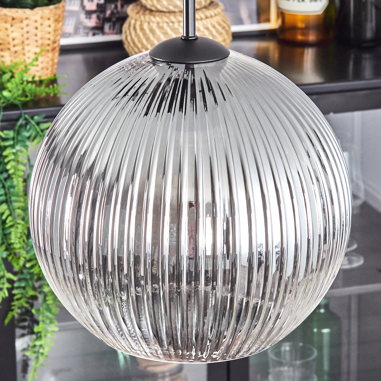 Eine klare, geriffelte Glaskugel-Pendelleuchte hängt. Der Hintergrund umfasst ein Regal mit Dekoration und Grün. Das Licht reflektiert die Umgebung.