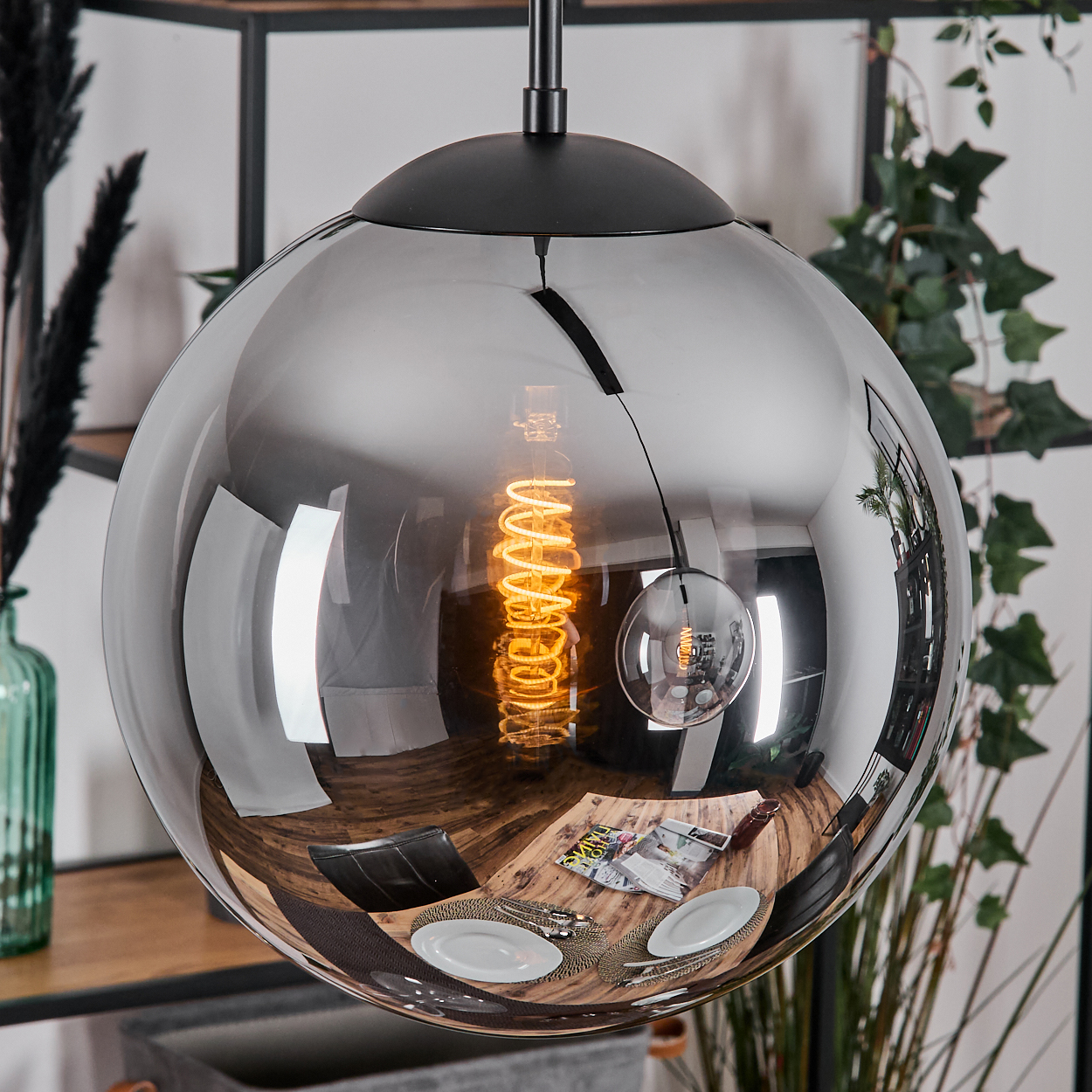 Eine moderne Pendelleuchte mit reflektierendem Kugelschirm. Im Inneren leuchtet eine Wendelglühbirne. Die Reflexion zeigt einen Esstisch und Pflanzen.