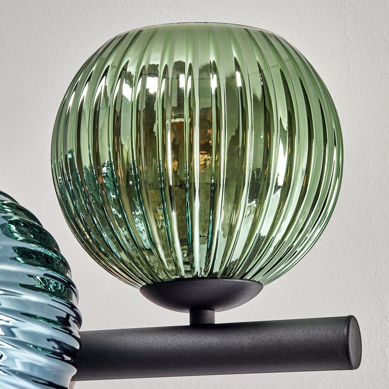 Nahaufnahme einer grünen, gerippten Glaslampe. Sie ist an einer schwarzen Metallstange an einer weißen Wand befestigt. Eine andere blaue Glaslampe ist in der Nähe.