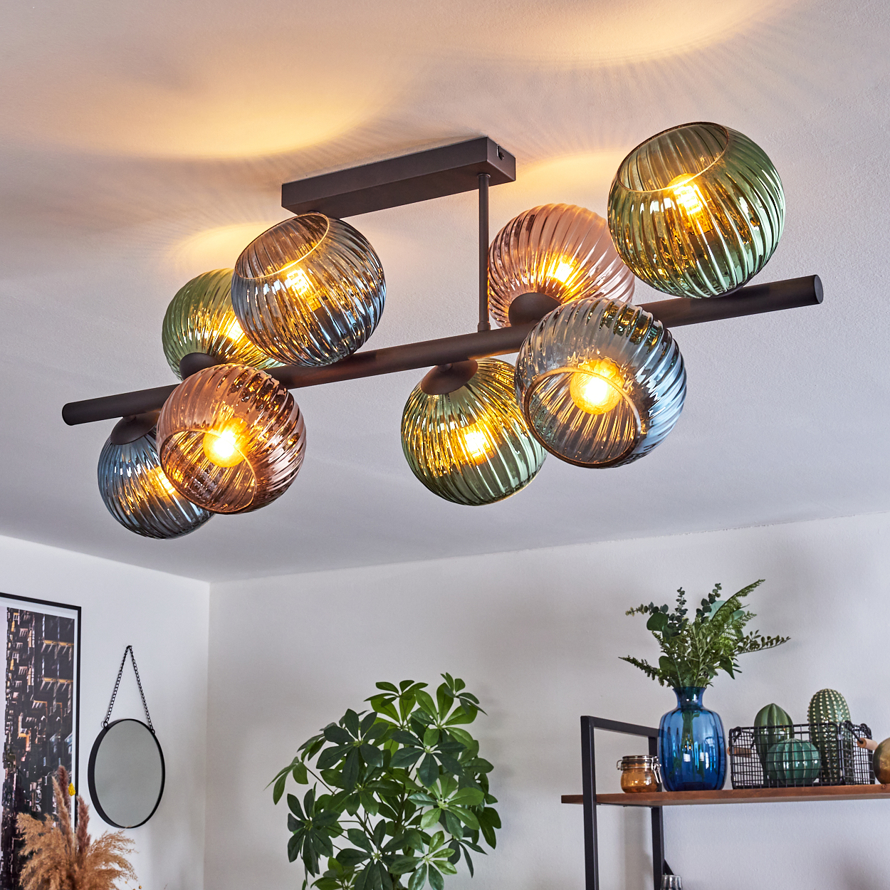Eine moderne Deckenleuchte mit acht Glaskugeln in verschiedenen Farben. Das Licht ist an und beleuchtet den Raum. Pflanzen und Dekoration sind im Hintergrund.