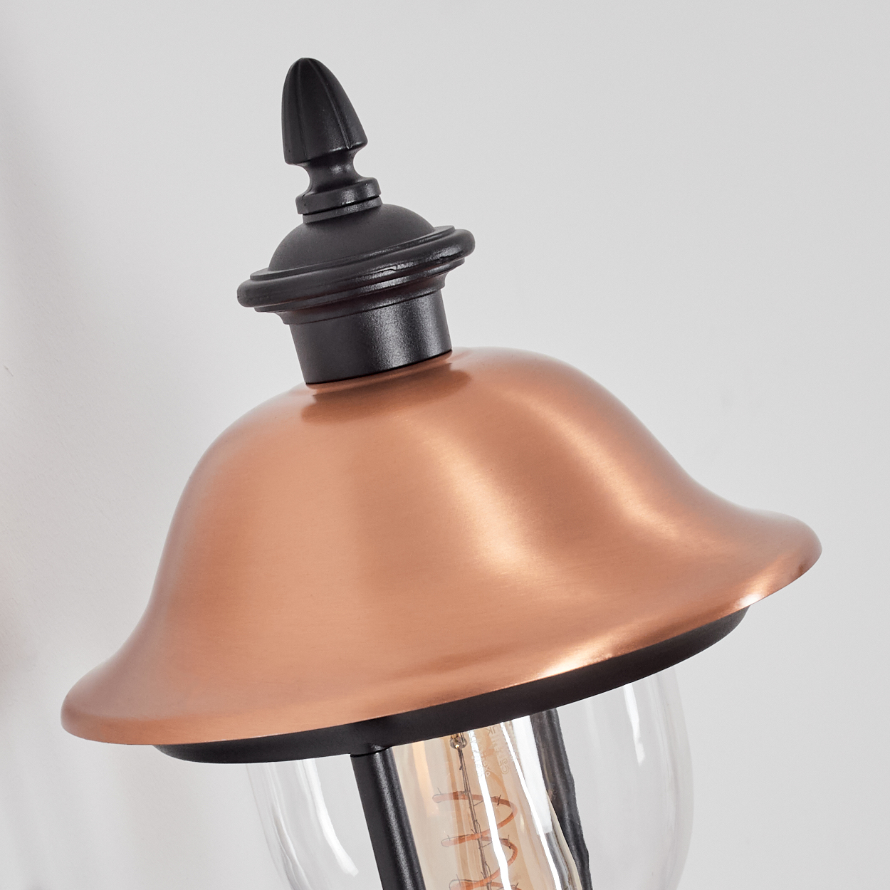 Nahaufnahme einer kupferfarbenen Außenlampe. Sie hat einen schwarzen Deckel und eine klare Glaskugel. Der Hintergrund ist weiß.