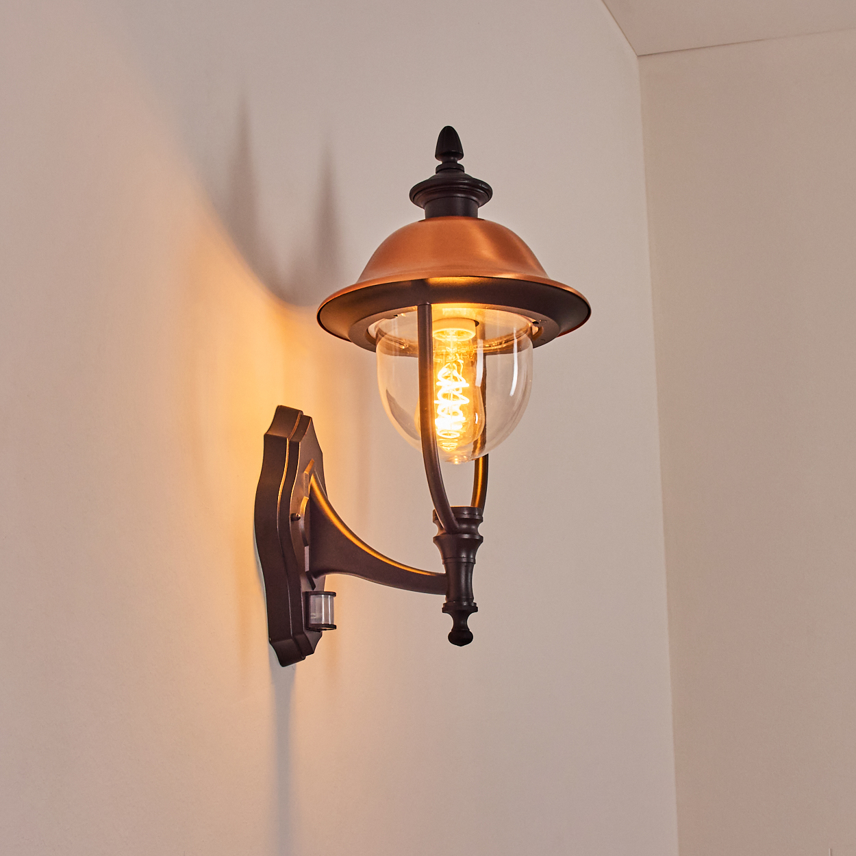 Eine Wand-Außenlampe mit kupferfarbenem Schirm und Glaskolben. Die Lampe ist an und wirft ein warmes Licht an die Wand.