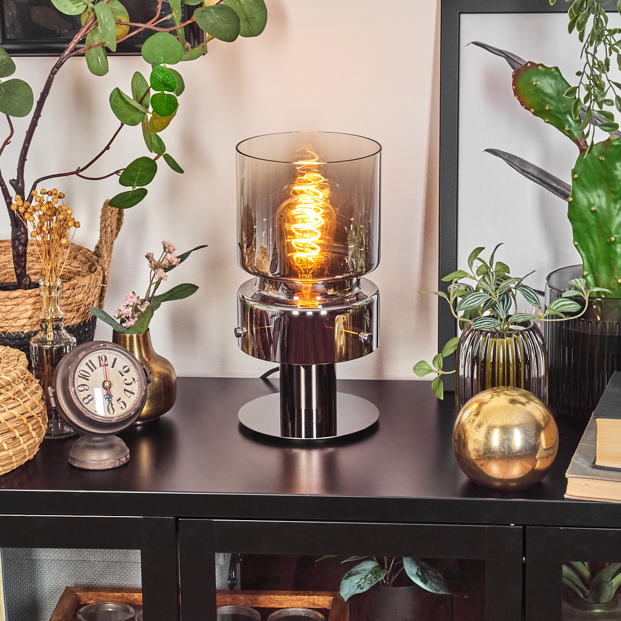 Eine beleuchtete Lampe auf einer schwarzen Oberfläche. Pflanzen, eine Uhr und eine goldene Kugel sind darum herum. Der Hintergrund hat ein Bild mit Rahmen.