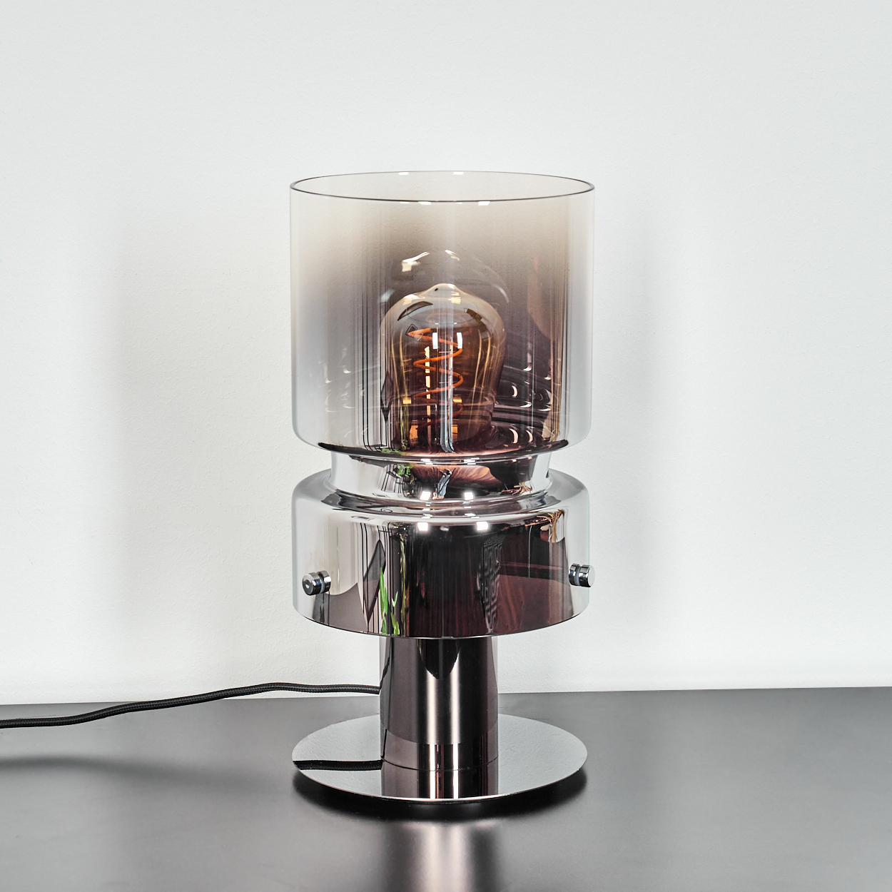 Eine moderne Chromlampe mit klarem Glasschirm, in dem sich eine Glühbirne im Vintage-Stil befindet. Sie steht auf dunkler Oberfläche vor weißer Wand.
