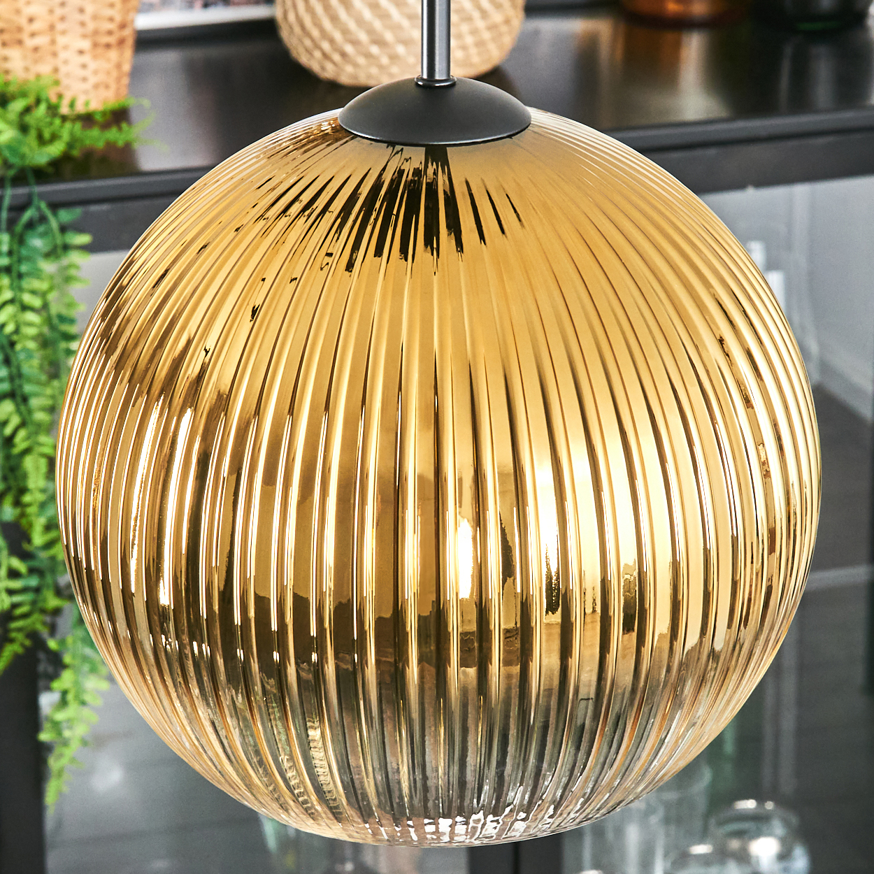 Eine goldene, gerippte Glas-Pendelleuchte hängt von der Decke. Grüne Pflanzen und ein geflochtener Korb sind im Hintergrund.