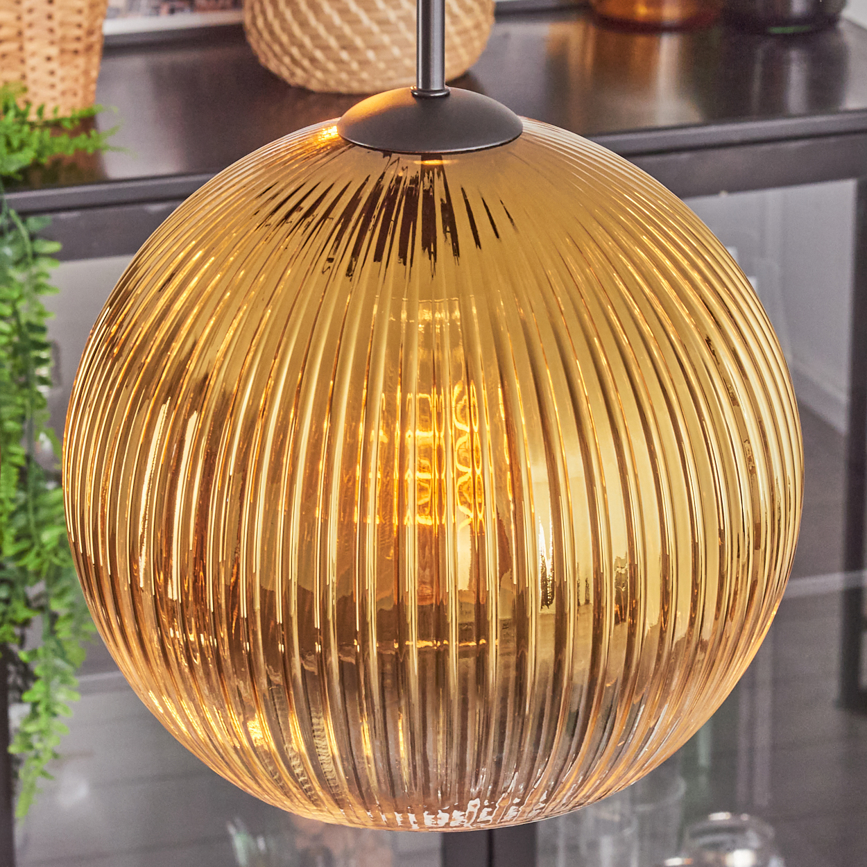 Eine goldene, gerippte Glas-Pendelleuchte hängt. Die Glühbirne leuchtet im Inneren. Ein Korb und grünes Laub sind im Hintergrund.