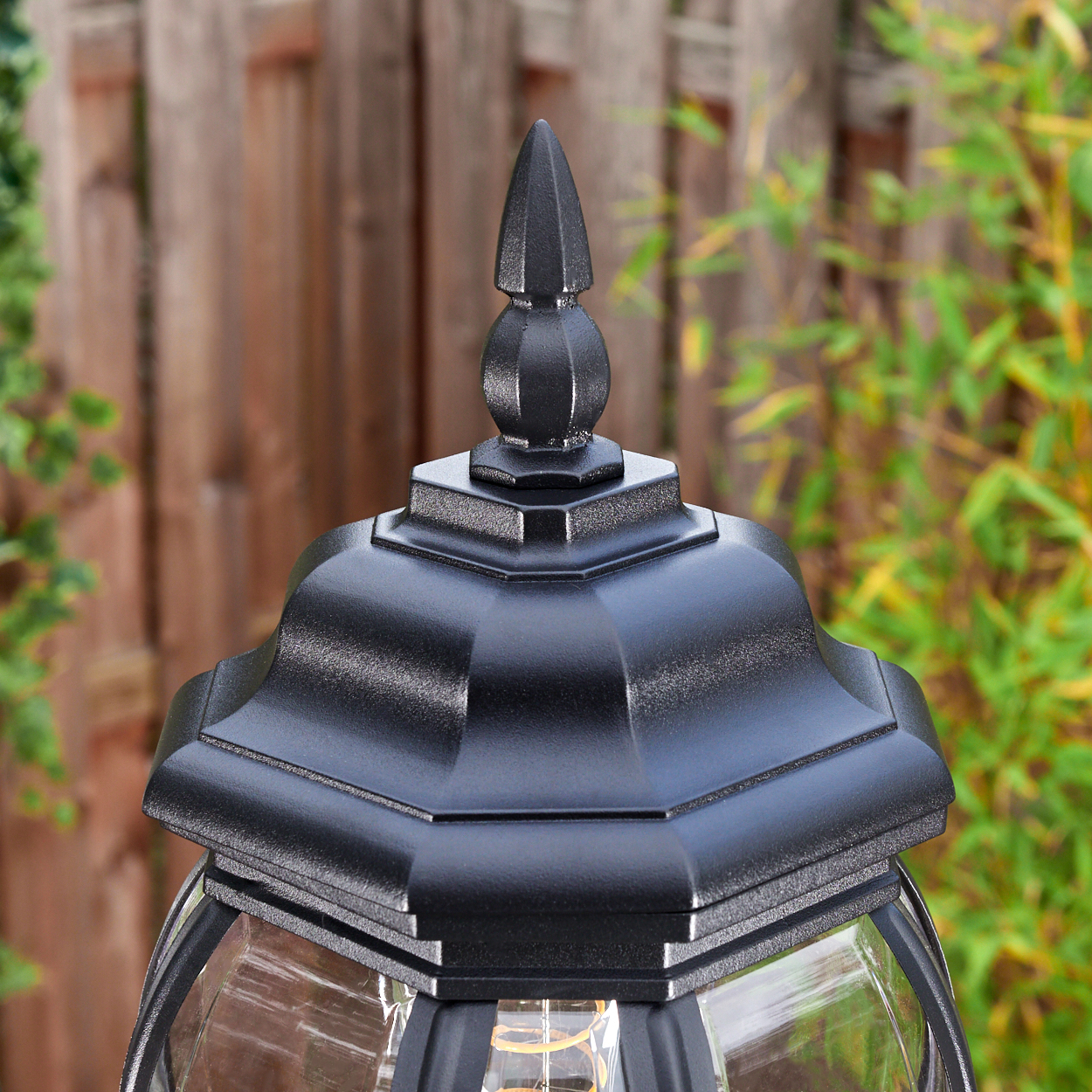 Nahaufnahme einer schwarzen Außenlampe mit spitzer Spitze. Im Hintergrund ein Holzzaun und grüne Pflanzen.