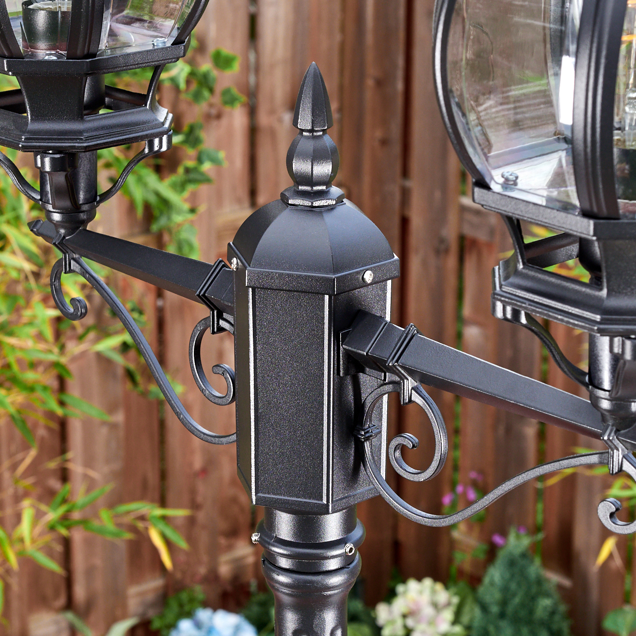 Eine schwarze Laterne mit zwei Lampen. Der Hintergrund umfasst einen Holzzaun und grüne Pflanzen.