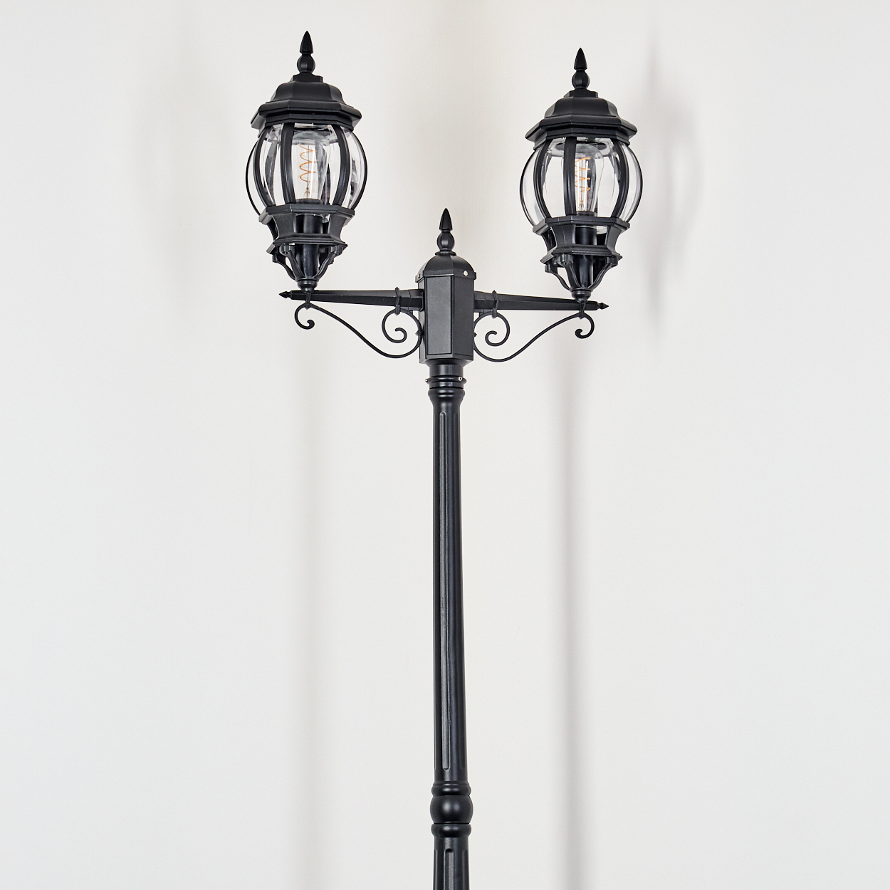 Eine schwarze, verzierte Straßenlaterne mit zwei Kugeln vor einer schlichten weißen Wand.
