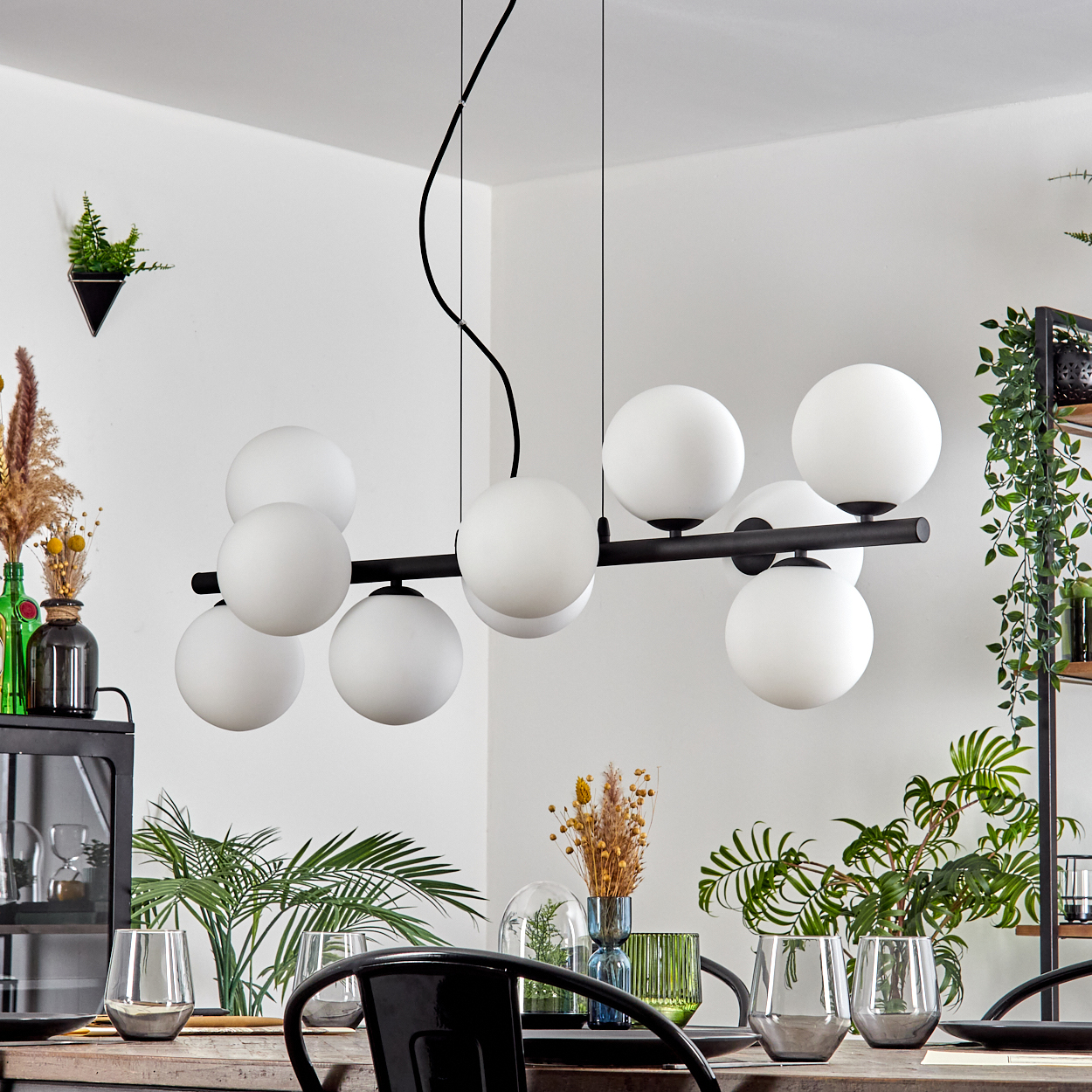 Ein modernes Esszimmer mit einem schwarzen Kronleuchter mit weißen Kugeln, Pflanzen, Gläsern und einem Holztisch.