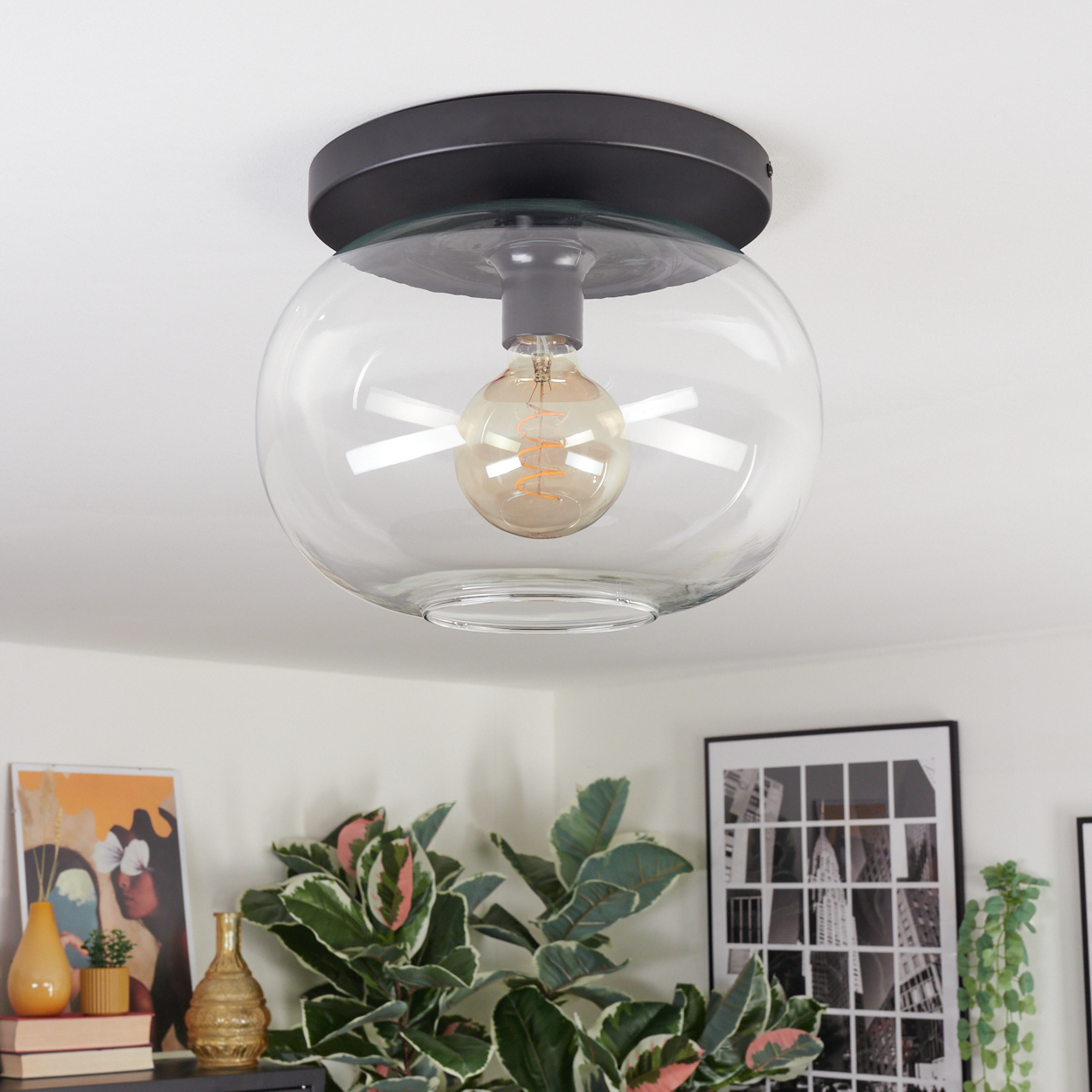 Eine moderne Deckenlampe mit Glaskugel. Im Inneren befindet sich eine leuchtende Glühbirne, im Hintergrund grüne Pflanzen und Kunstwerke.
