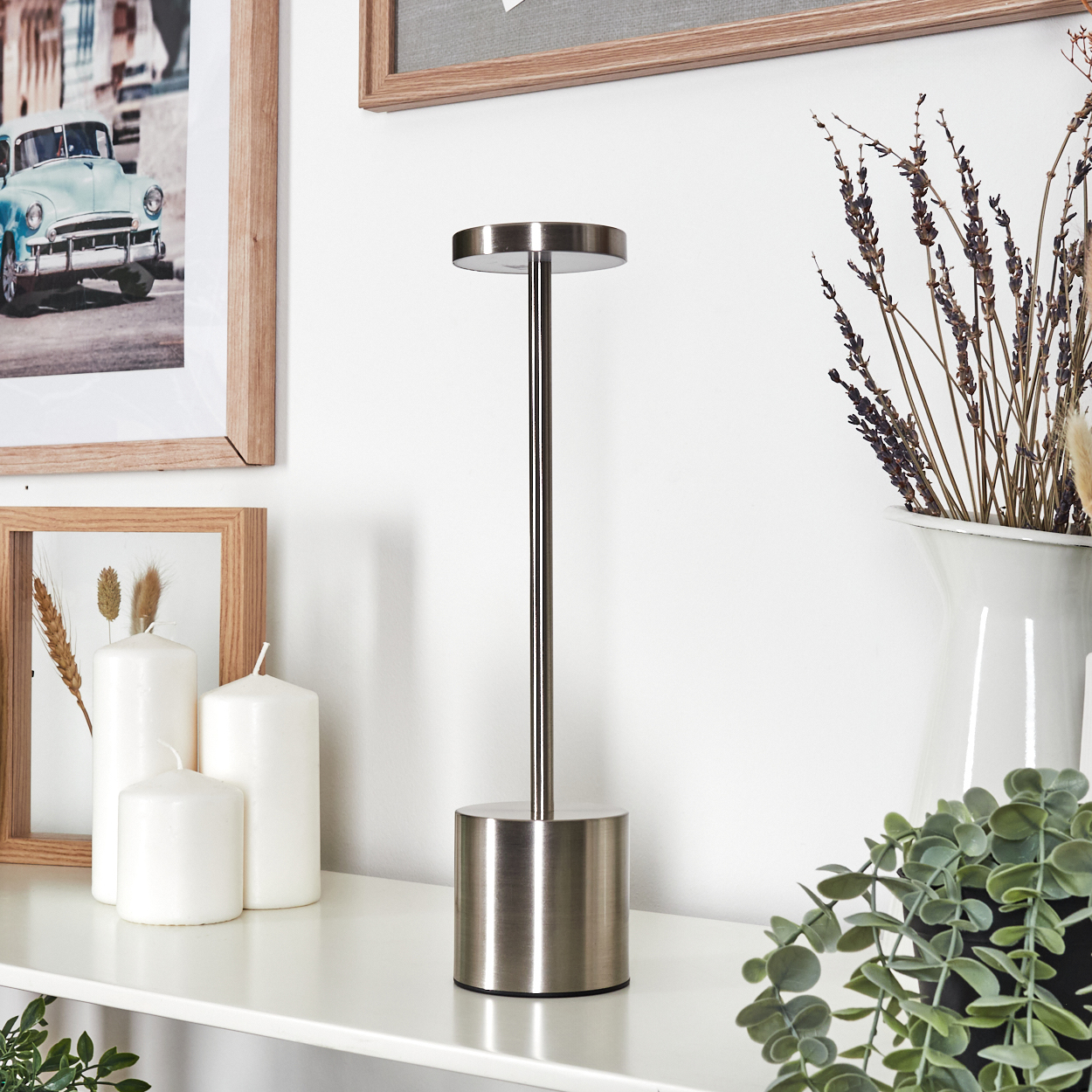 Eine moderne Lampe steht auf einem weißen Regal mit Kerzen und gerahmten Bildern. Trockenblumen in einer Vase und Grünzeug sind ebenfalls vorhanden.