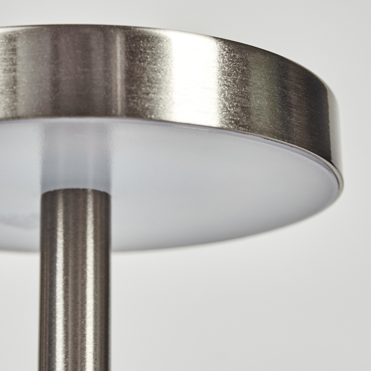 Nahaufnahme einer Lampe. Metallischer zylindrischer Stiel trägt einen runden, gebürsteten Metallschirm mit weißer Oberfläche.