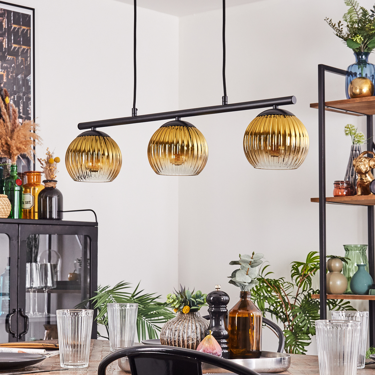 Esszimmer mit gedecktem Tisch. Drei goldene Pendelleuchten hängen darüber. Regale mit Vasen und Dekor sind sichtbar.