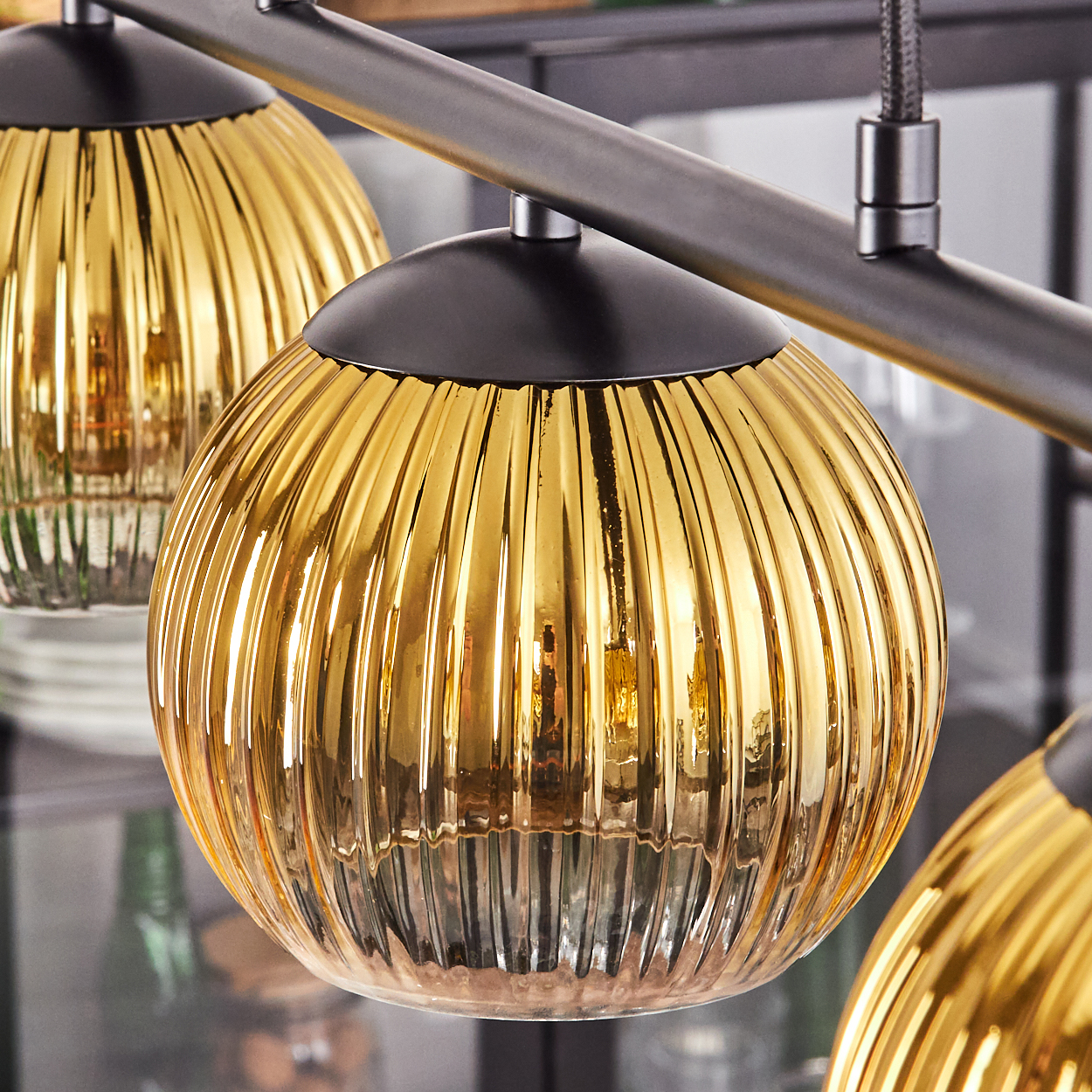 Nahaufnahme eines modernen Kronleuchters. Drei goldfarbene Glaskugeln mit vertikalen Rippen hängen an einem dunklen Metallrahmen.