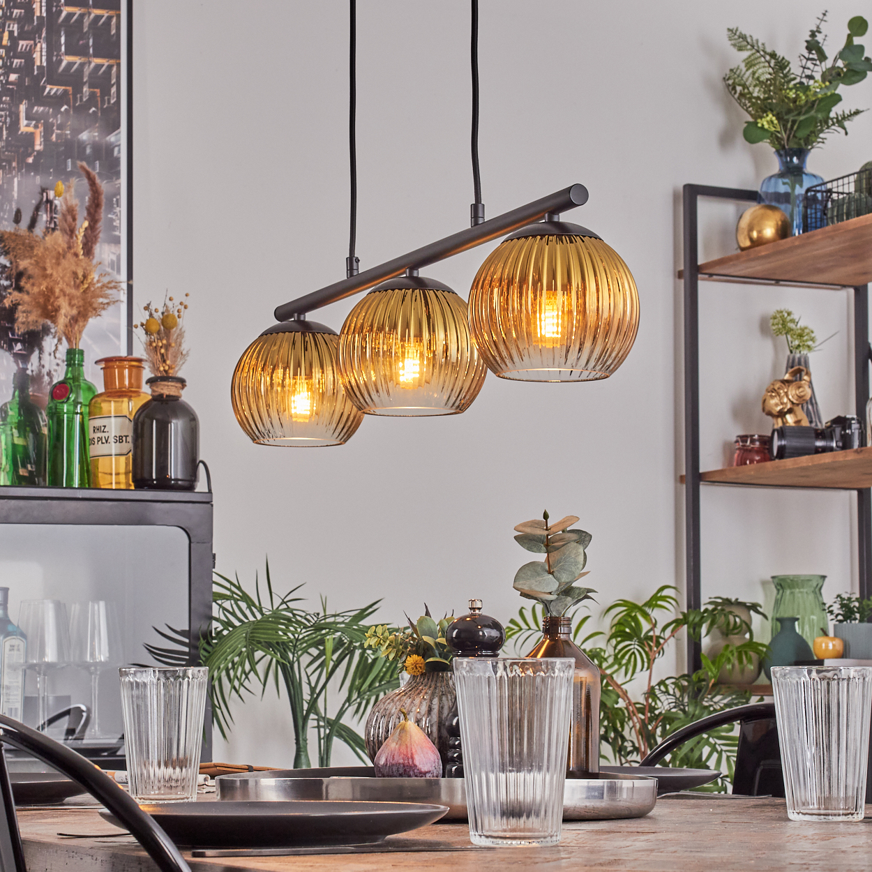 Esszimmer mit Hängelampen, Tischarrangement und Pflanzen. Drei Lampen hängen über einem Tisch mit Gläsern und Geschirr.