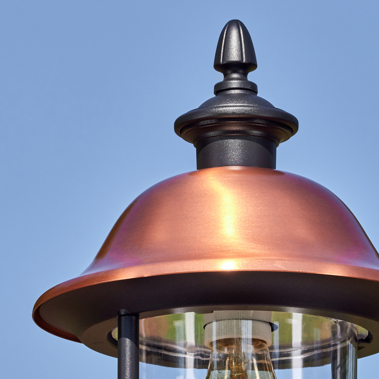 Nahaufnahme einer Kupferlampe mit schwarzem Oberteil vor blauem Himmel. Die Lampe hat ein Glasgehäuse und eine sichtbare Glühbirne.