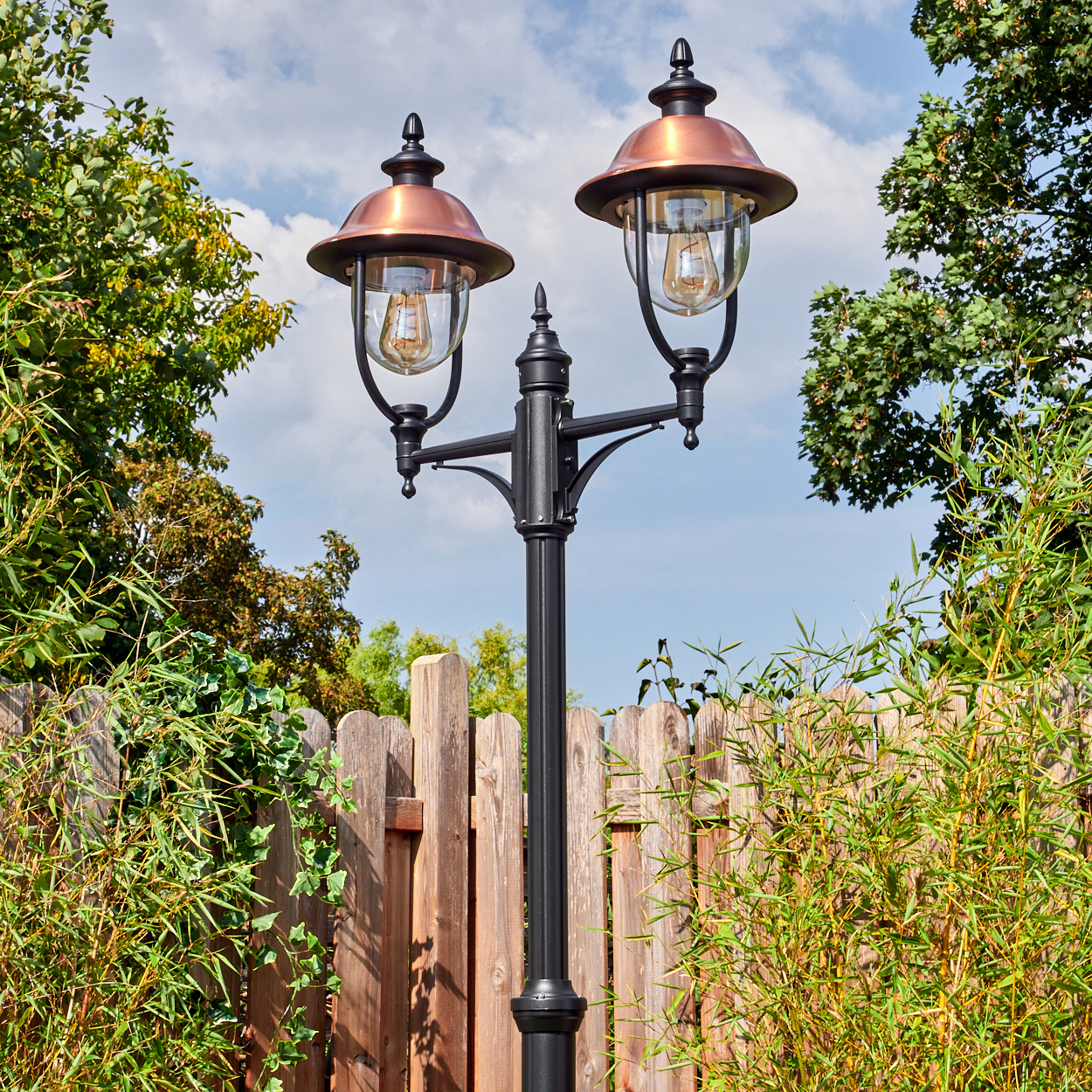 Schwarze Laterne mit zwei Lampen. Kupferaufsätze und Glasschirme. Holzzaun und grüne Pflanzen umgeben sie. Blauer Himmel im Hintergrund.