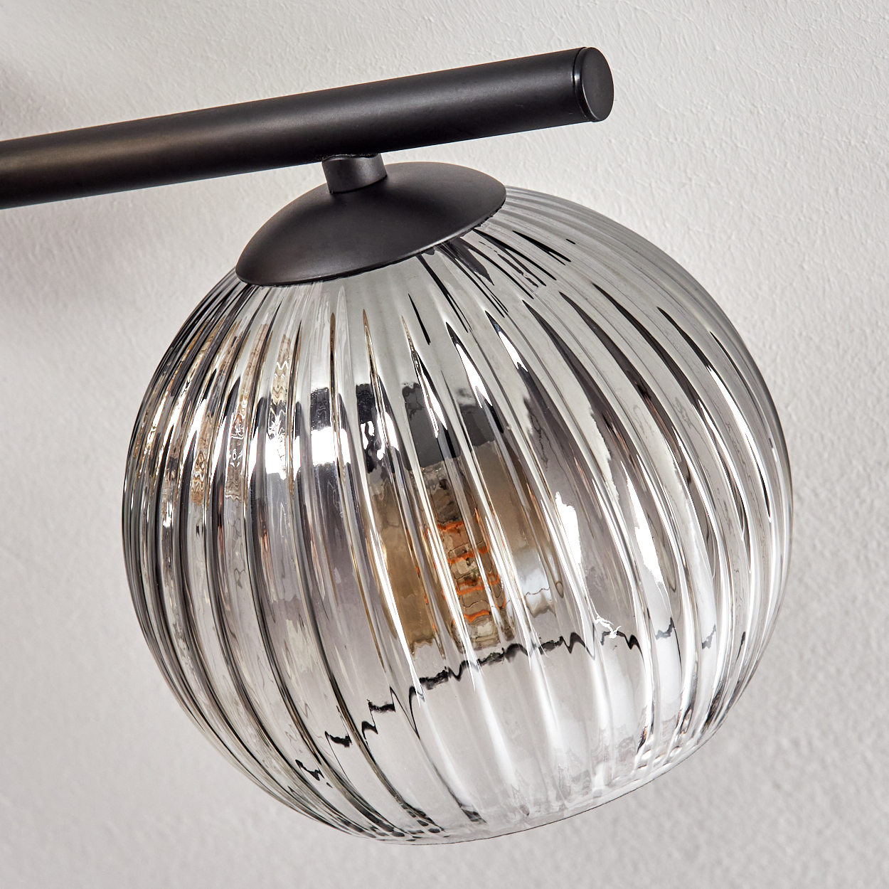 Nahaufnahme einer silbernen Glaskugel-Leuchte mit vertikalen Rippen. Sie ist an einem schwarzen Metallarm an einer weißen Wand befestigt.
