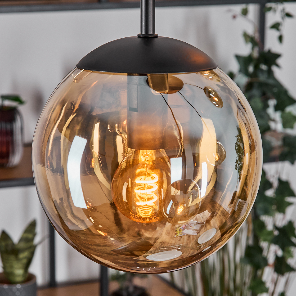 Eine runde, bernsteinfarbene Pendelleuchte. Eine leuchtende Wendelglühbirne befindet sich im Inneren. Die Lampe hängt von der Decke. Grüne Pflanzen im Hintergrund.