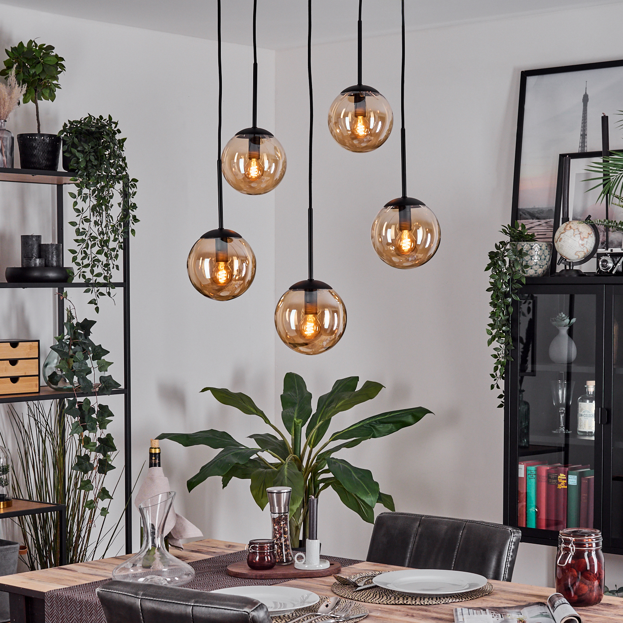 Esszimmer mit hängenden Kugelleuchten, einem Holztisch mit Tellern und einer großen Pflanze. Regale mit Dekoration im Hintergrund.