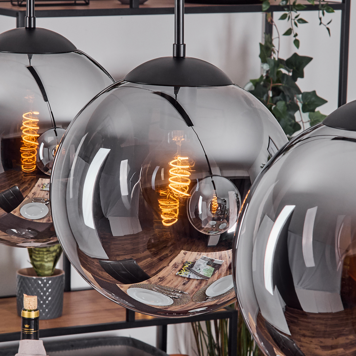 Drei kugelförmige Pendelleuchten. Jede hat ein Wendelfilament. Reflektiert Esstisch, Teller, Pflanzen im Hintergrund.