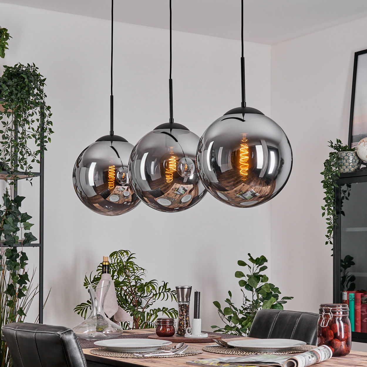 Drei kugelförmige Pendelleuchten hängen über einem Esstisch. Die Lampen sind silbern. Der Tisch ist mit Tellern gedeckt und es gibt Pflanzen.