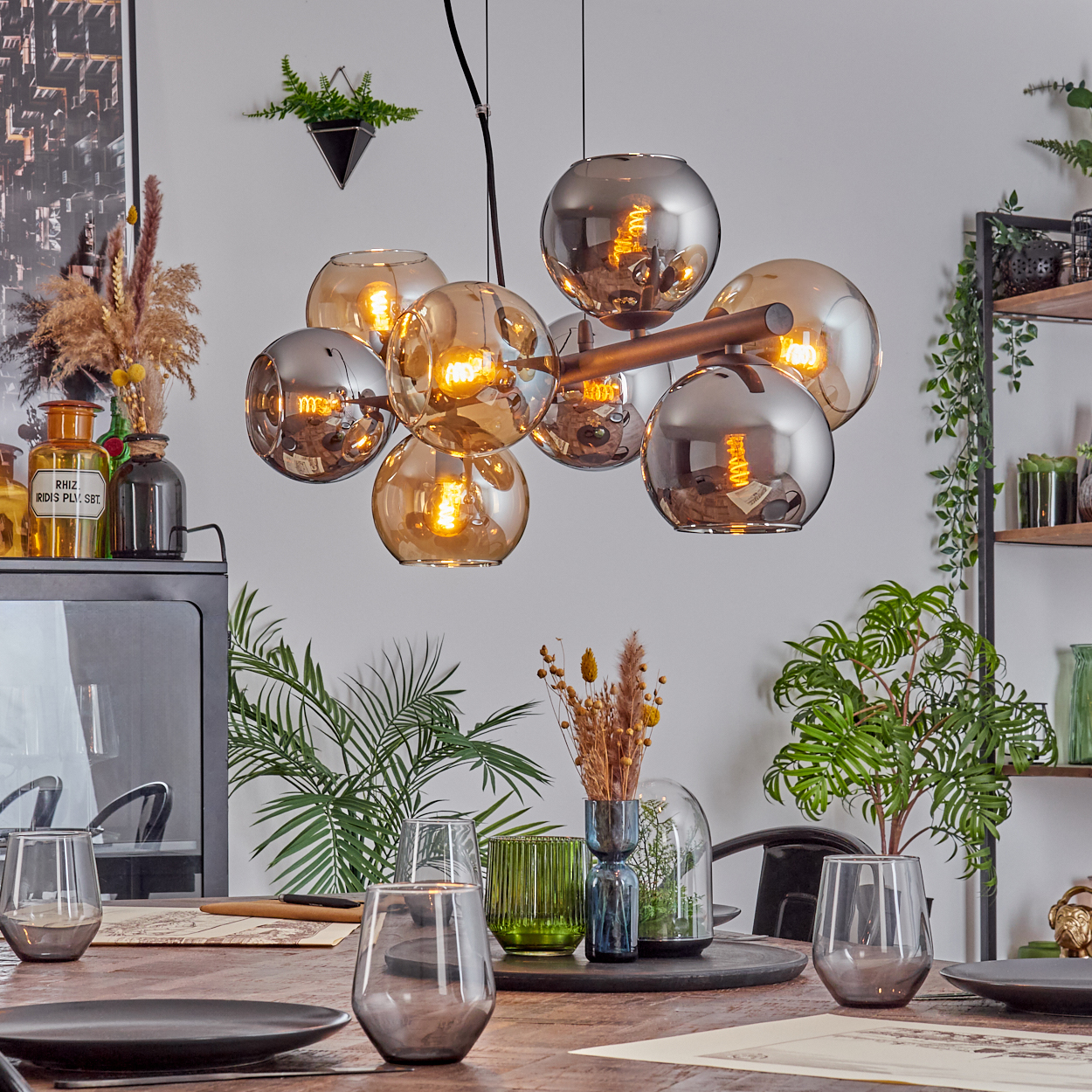 Esszimmer mit Holztisch, Gläsern, Pflanzen und einem modernen Kronleuchter mit kugelförmigen Lampen. Dekorative Gegenstände und Regale sind sichtbar.