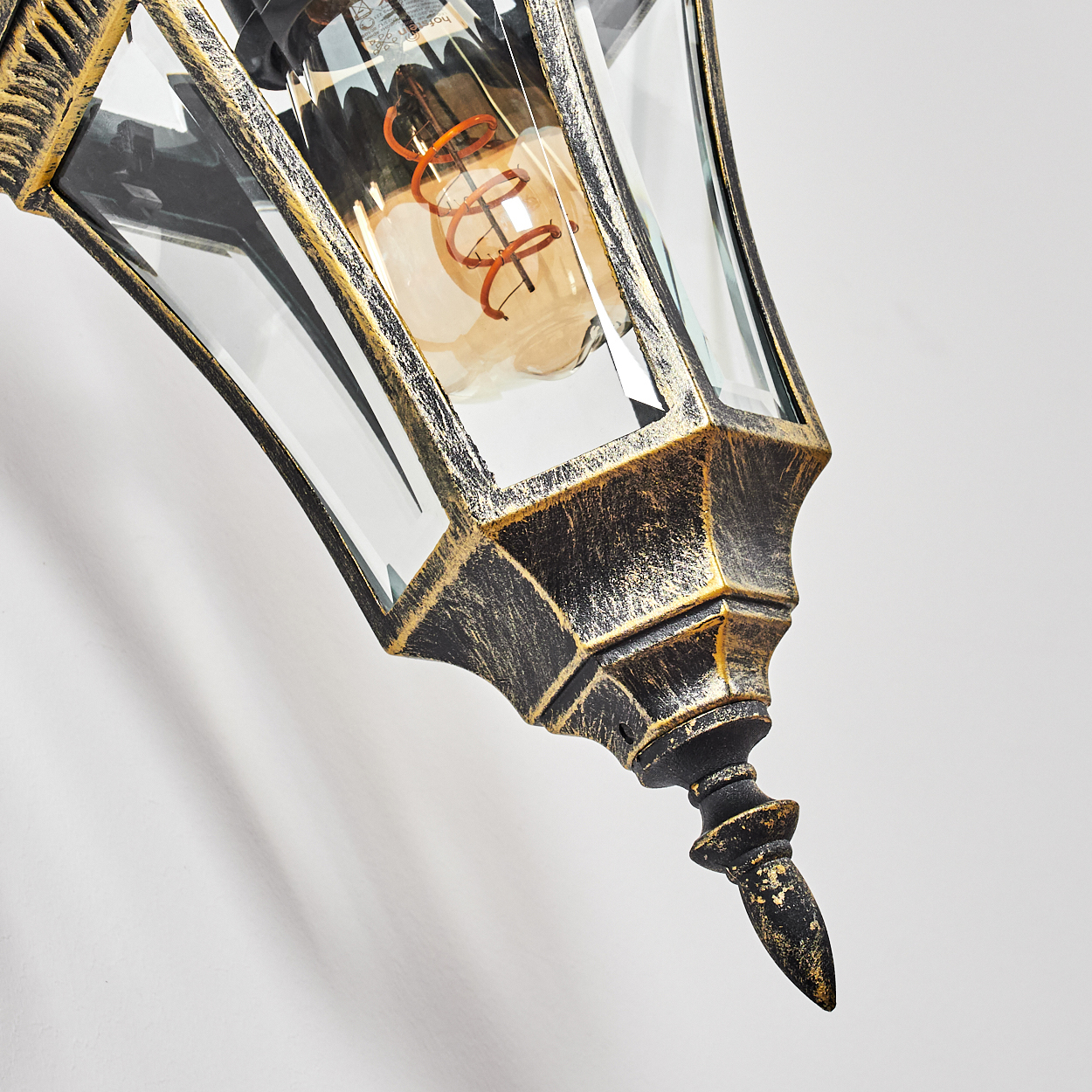 Nahaufnahme einer dekorativen Wandlampe mit gold-schwarzem Finish, Glaspaneelen und einer sichtbaren Glühbirne auf weißem Hintergrund.