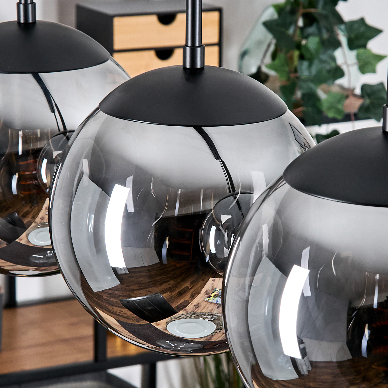 Drei kugelförmige, graue Glas-Pendelleuchten mit schwarzen Oberteilen. Im Hintergrund ein Holzschrank und eine grüne Pflanze.