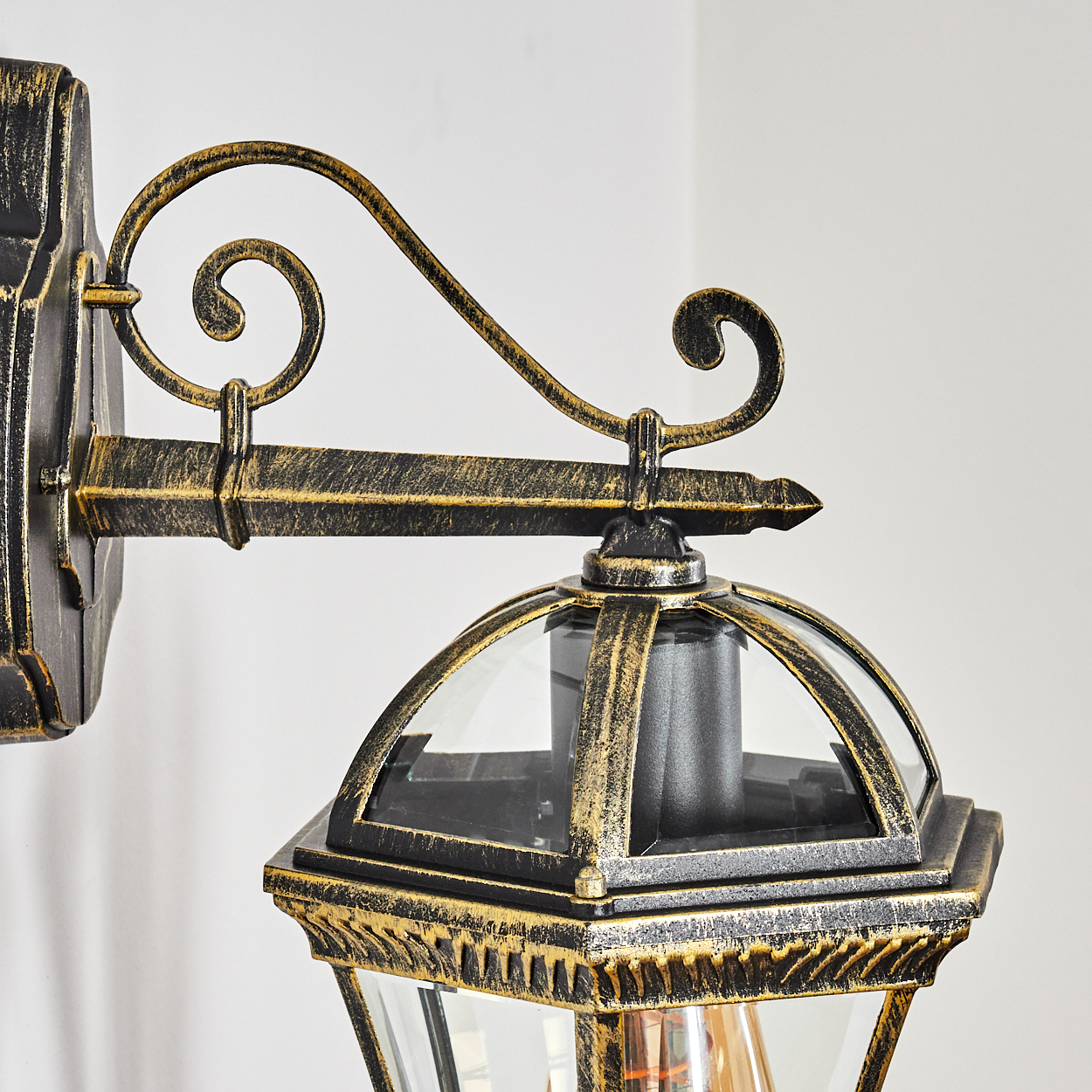 Nahaufnahme einer an der Wand montierten Außenlampe. Sie hat eine Bronzefinish, klare Glasscheiben und ein verziertes Design. Der Hintergrund ist weiß.