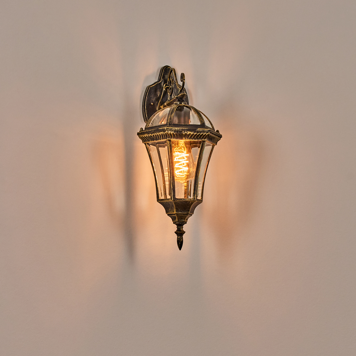 Eine verzierte, bronzefarbene Wandlampe mit einer leuchtenden Glühbirne, die einen Schatten auf eine hellfarbige Wand wirft.