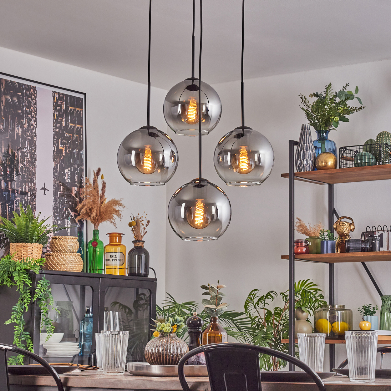 Esszimmer mit Hänge-Kugelleuchten, Pflanzen und einem schwarzen Tisch mit Stühlen. Regale und ein Bild vervollständigen die Szene.
