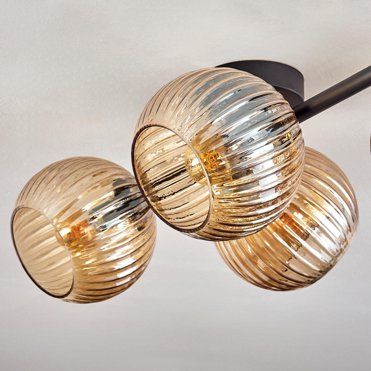 Eine moderne Deckenlampe mit drei bernsteinfarbenen Glaskugelschirmen, geripptem Design und schwarzen Metallarmen vor weißer Decke.