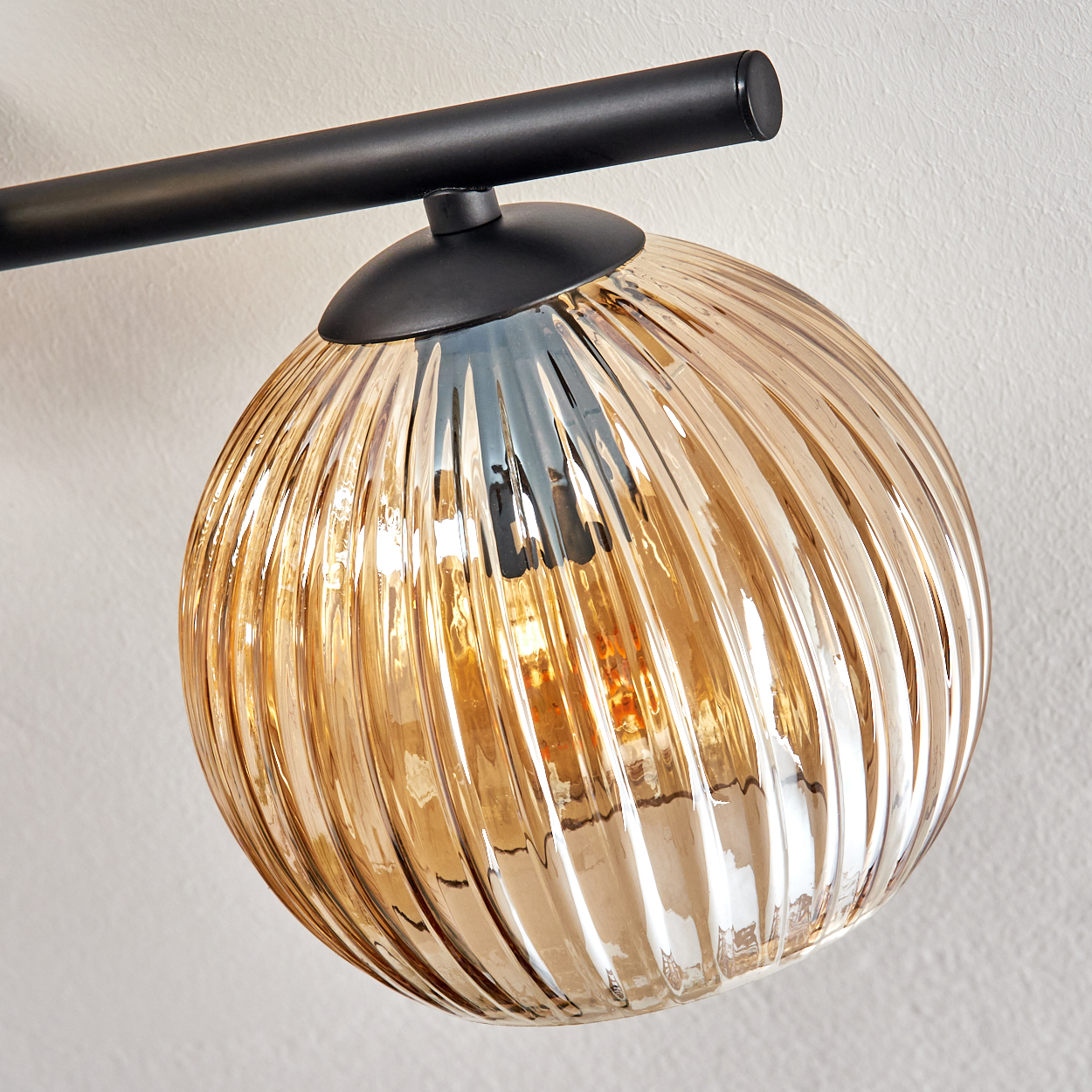 Nahaufnahme einer stilvollen Lampe mit geripptem, goldenem Glasschirm. Sie hängt an einer schwarzen Metallhalterung an einer weißen Wand.